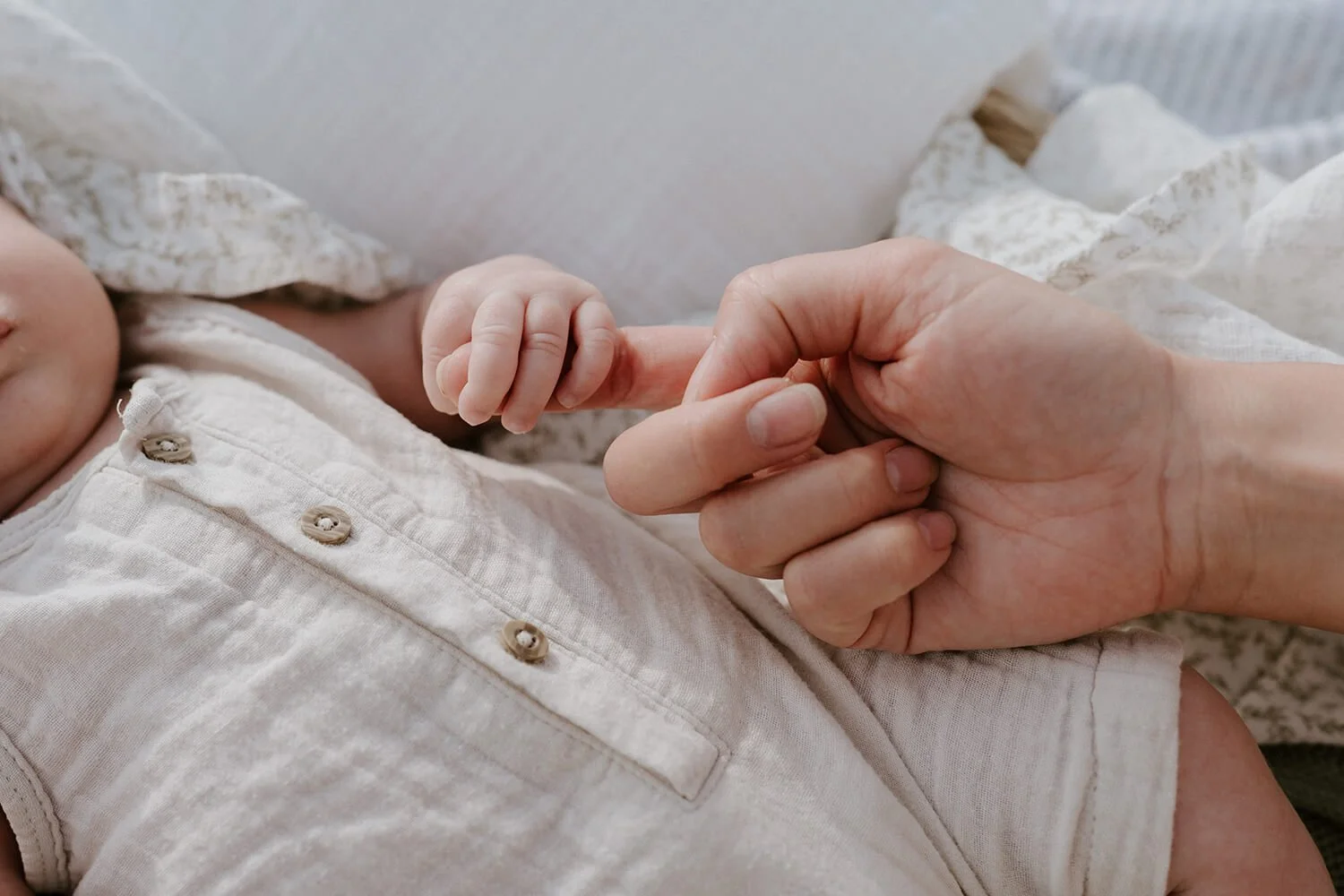 Close-up of an adult holding a baby's hand, with the baby's fingers wrapped around the adult's index finger, on a bed with a light-colored blanket and pajamas.