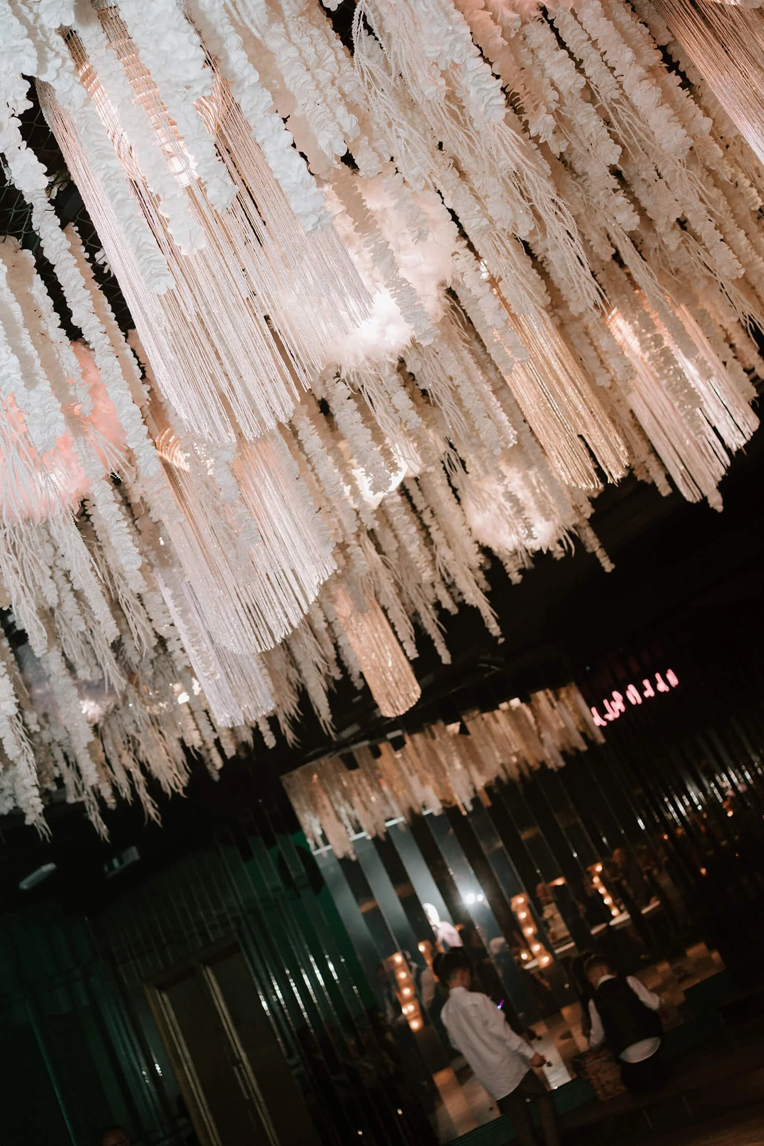 Elegant ceiling decor with hanging white and pink floral strands and ribbons, and softly lit with warm lights in a dark atmosphere. People are seen sitting and standing in the background of a stylish venue.