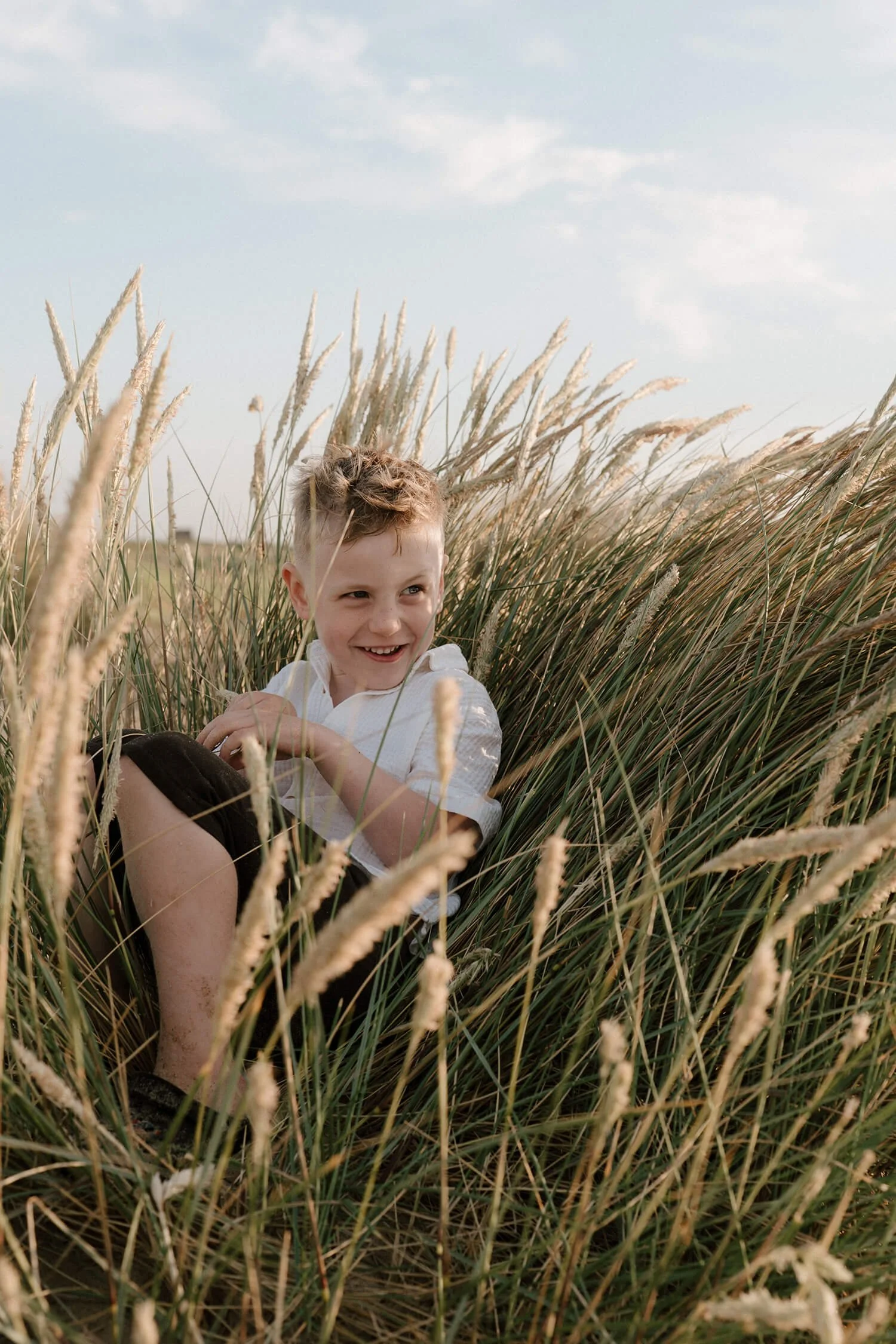 A young boy smiling and playing in tall, golden grass outdoors during the daytime under a partly cloudy sky.