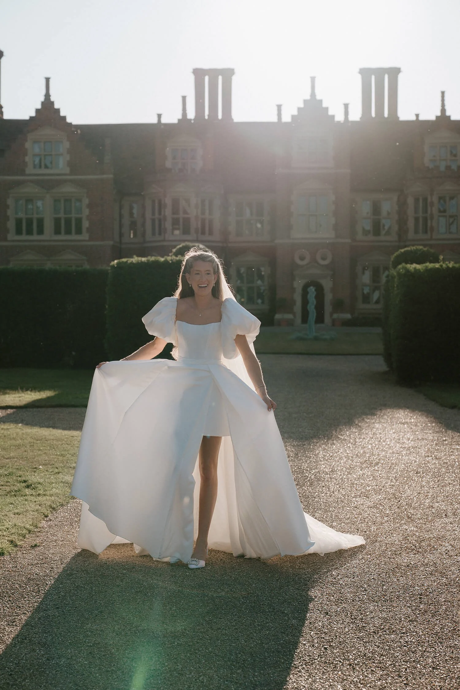A woman in a white wedding dress standing on a gravel path outside a large mansion, holding up the sides of her dress, smiling, with sunlight shining behind her.