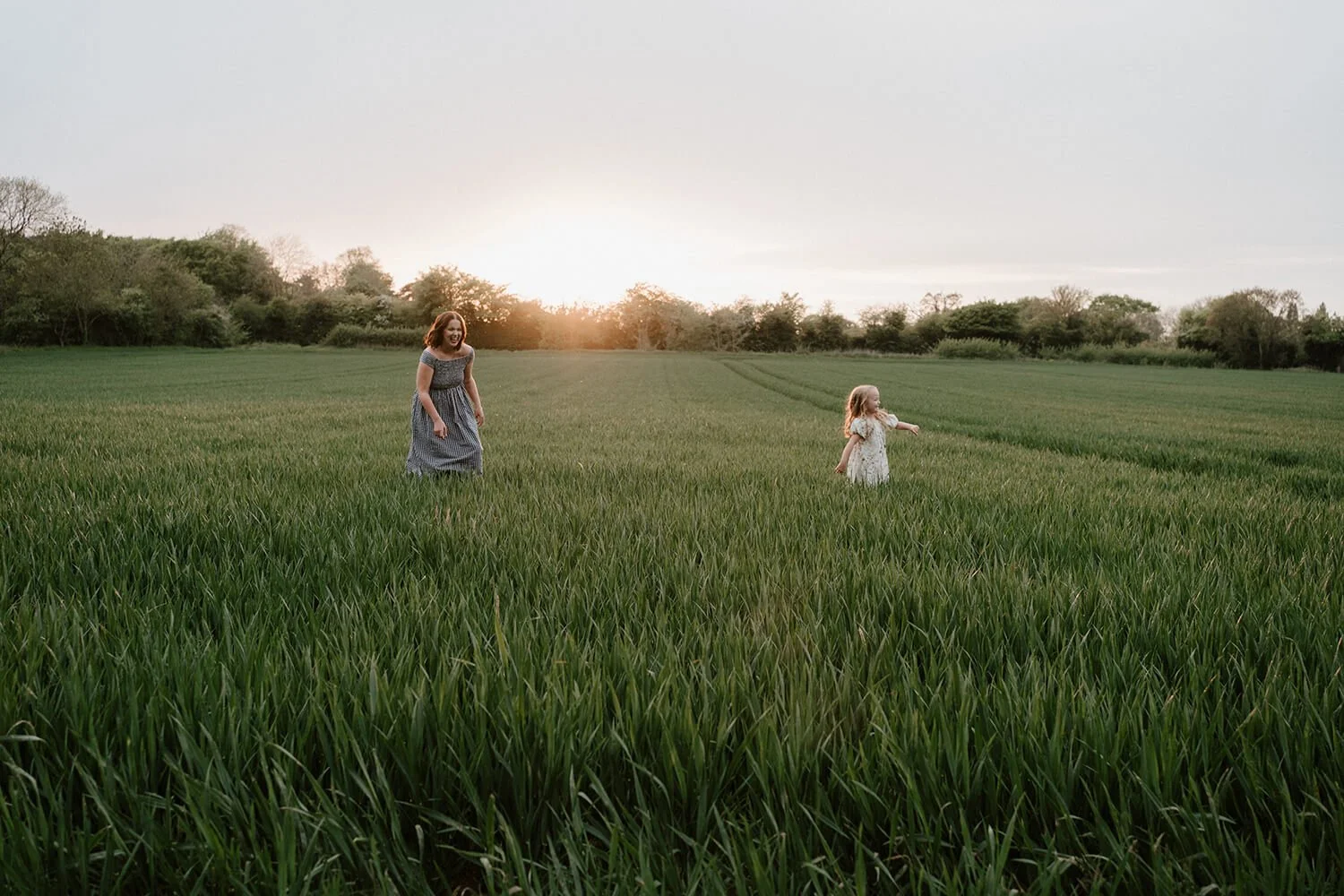A woman and a young girl walking through a lush green field at sunset, with trees in the background.