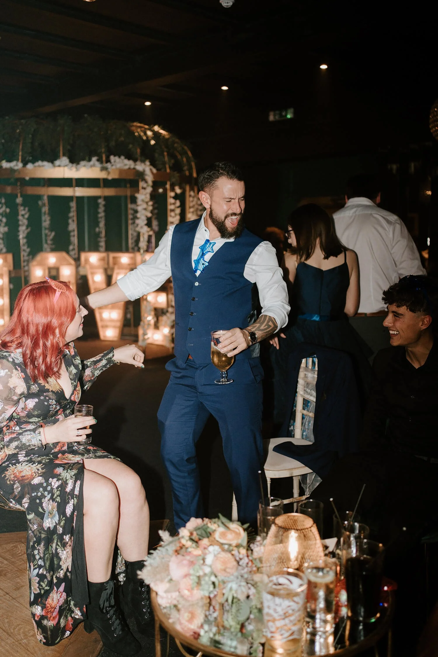 A man with a beard, wearing a blue vest and navy pants, is dancing and holding a drink at a celebration. A woman with red hair in a floral dress is sitting nearby, also holding a drink. The scene is lively with other people in the background, decorat