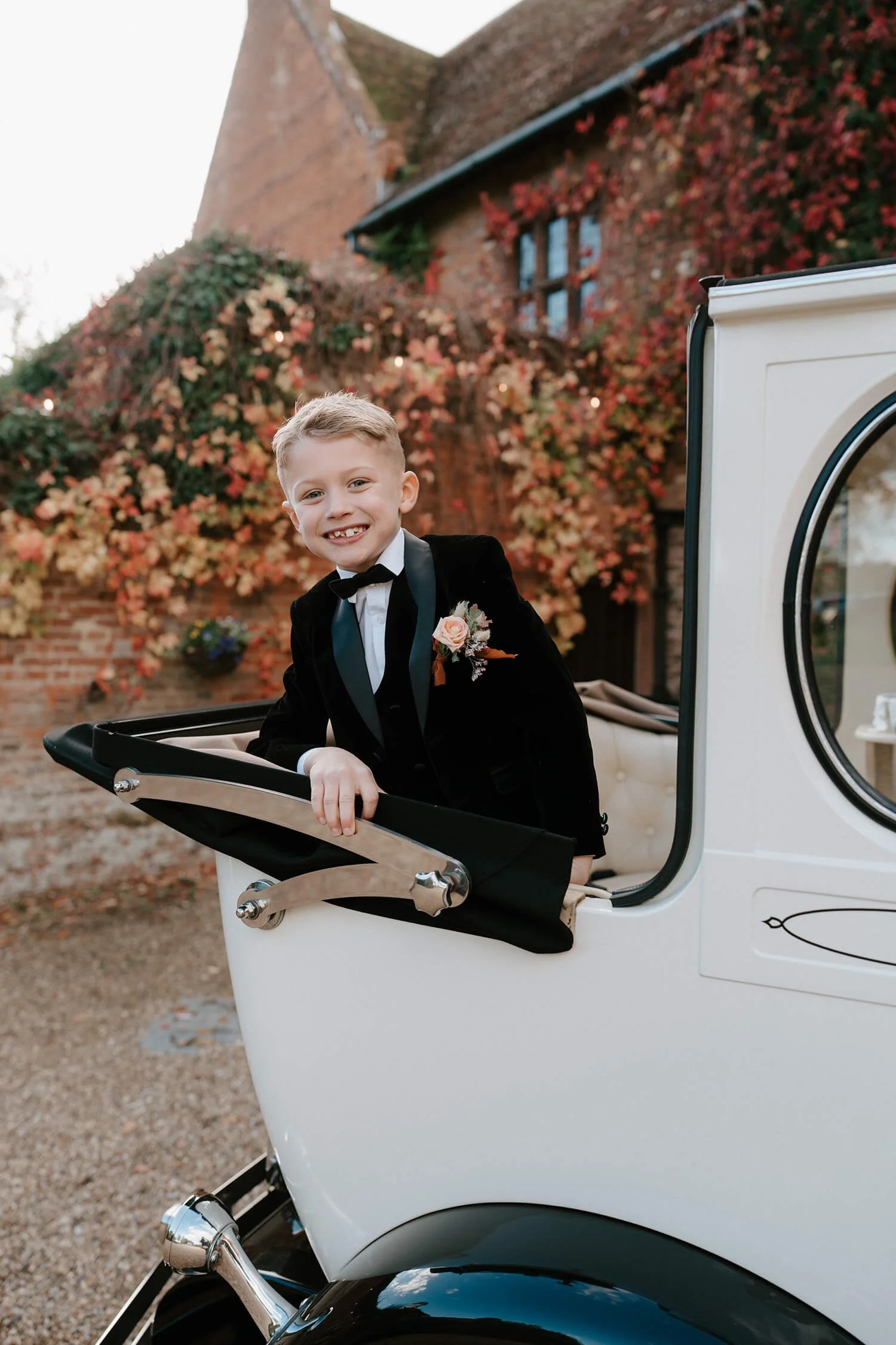 A young boy dressed in a tuxedo standing in the carriage of a vintage white car with a black interior, smiling at the camera, with a building covered in red and pink ivy in the background.
