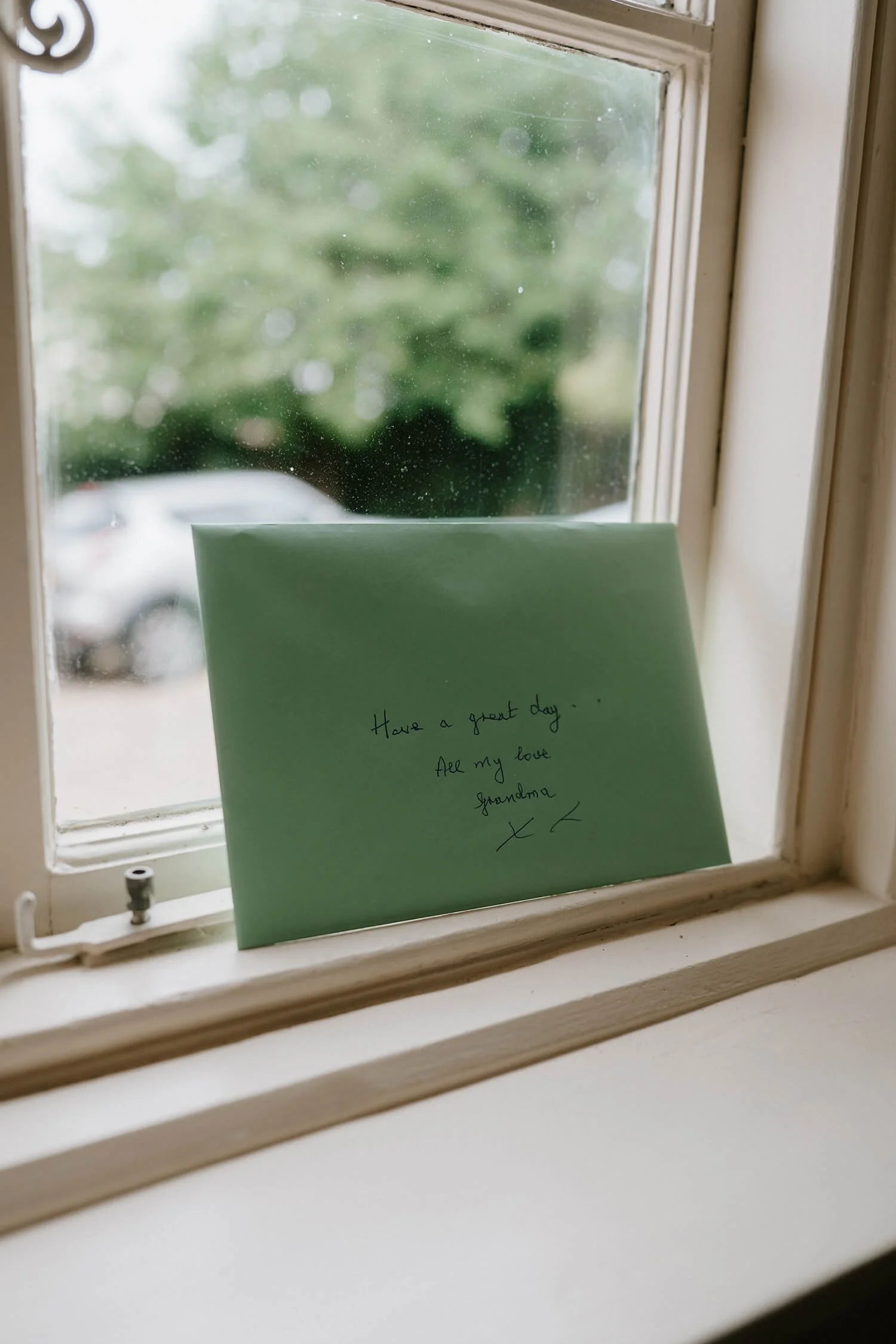A light green card with handwritten message placed on a windowsill. The message reads: 'Have a great day. All my love Grandma'. Outside, there is a blurred view of a parked car and greenery.