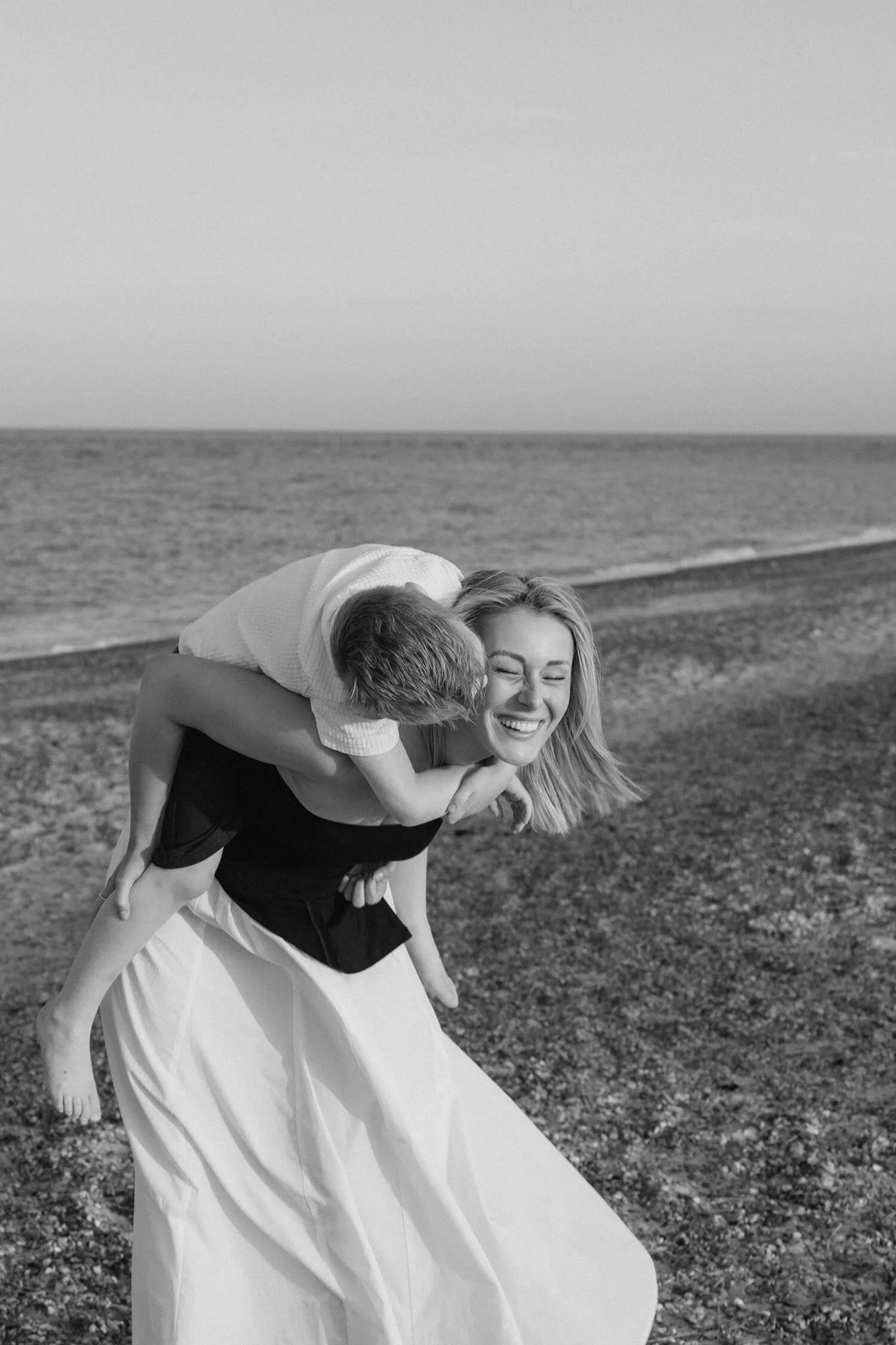 A woman at the beach is giving a piggyback ride to a young boy. They are both smiling and appear happy.