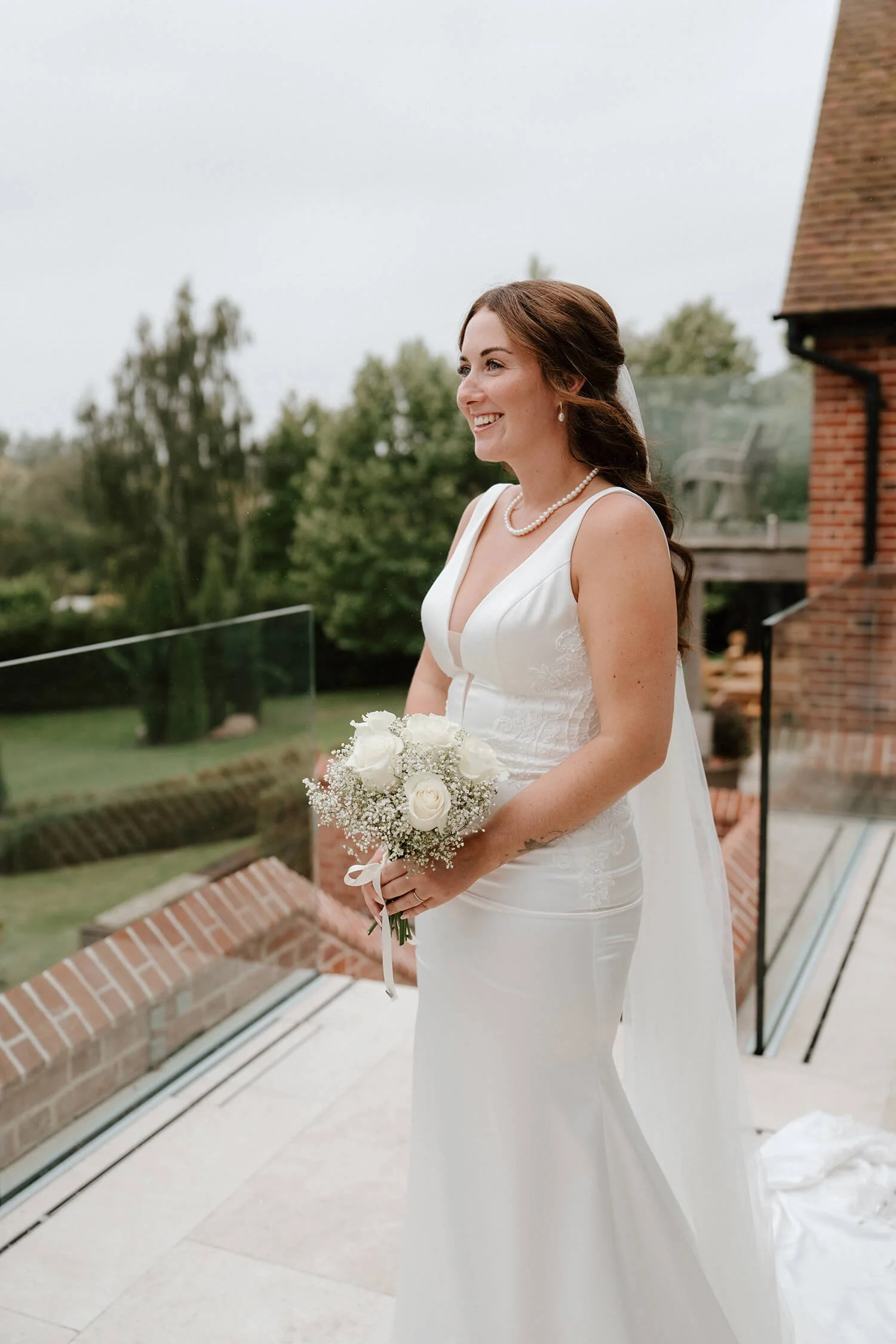 A smiling bride in a white wedding dress holding a bouquet of white roses and baby's breath on a balcony with a glass railing, outdoor greenery, and brick building in the background.