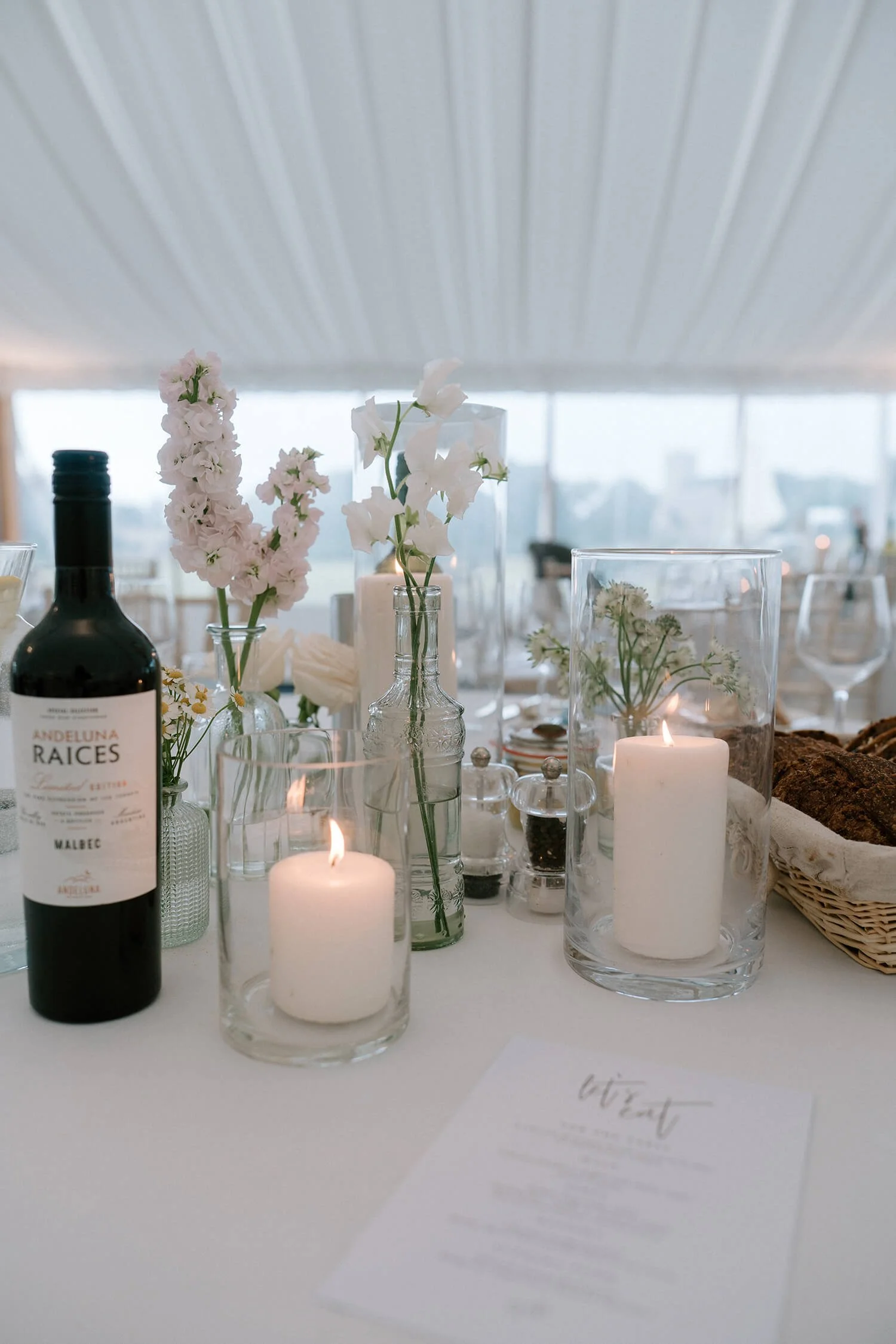 Decorative table setting with candles, flowers in glass bottles, a bottle of wine, and a basket of bread on a white tablecloth, in a well-lit room with large windows.