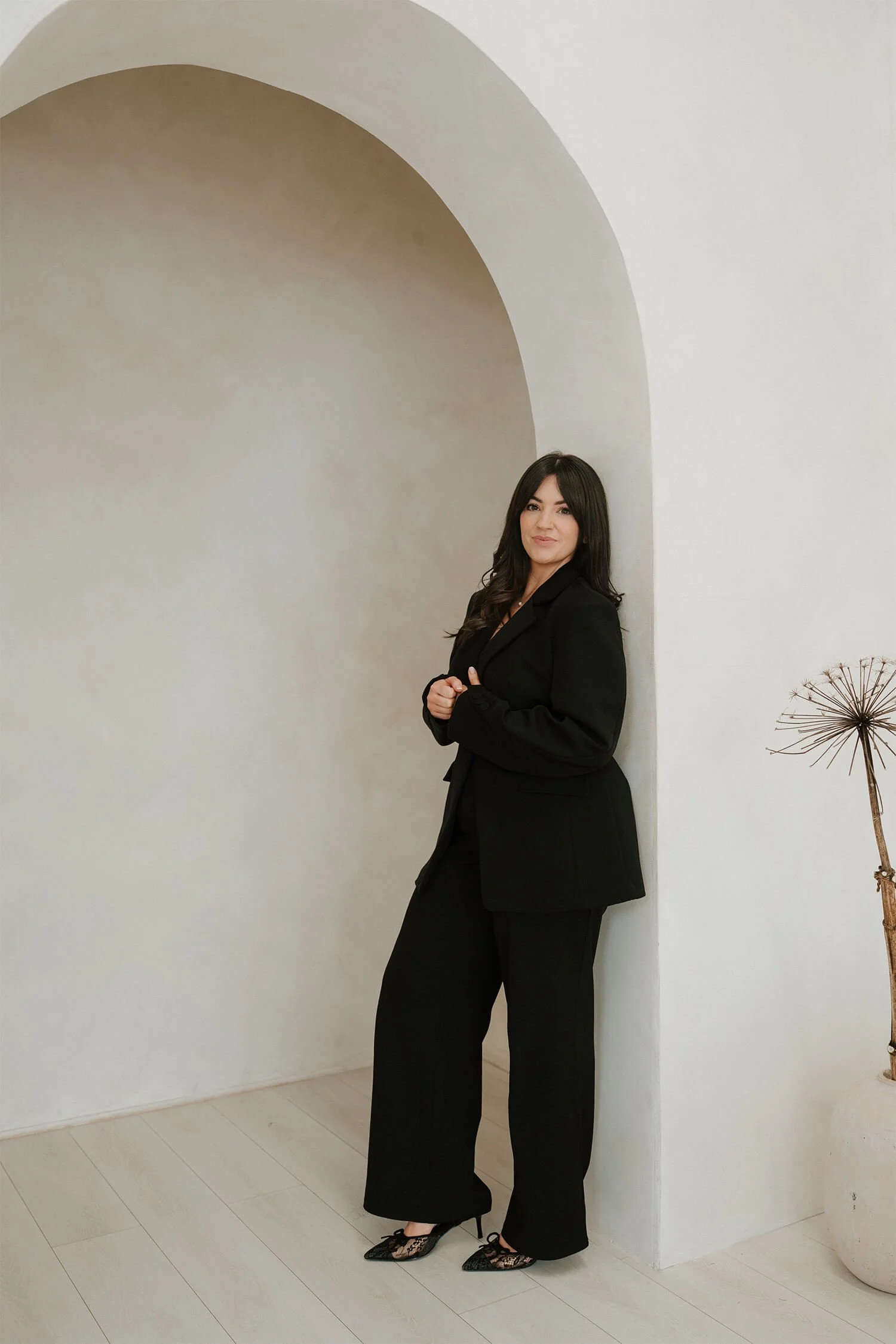 A woman in a black suit and high heels stands against a white wall inside a minimalist room with a curved archway and a dried plant in a pot.
