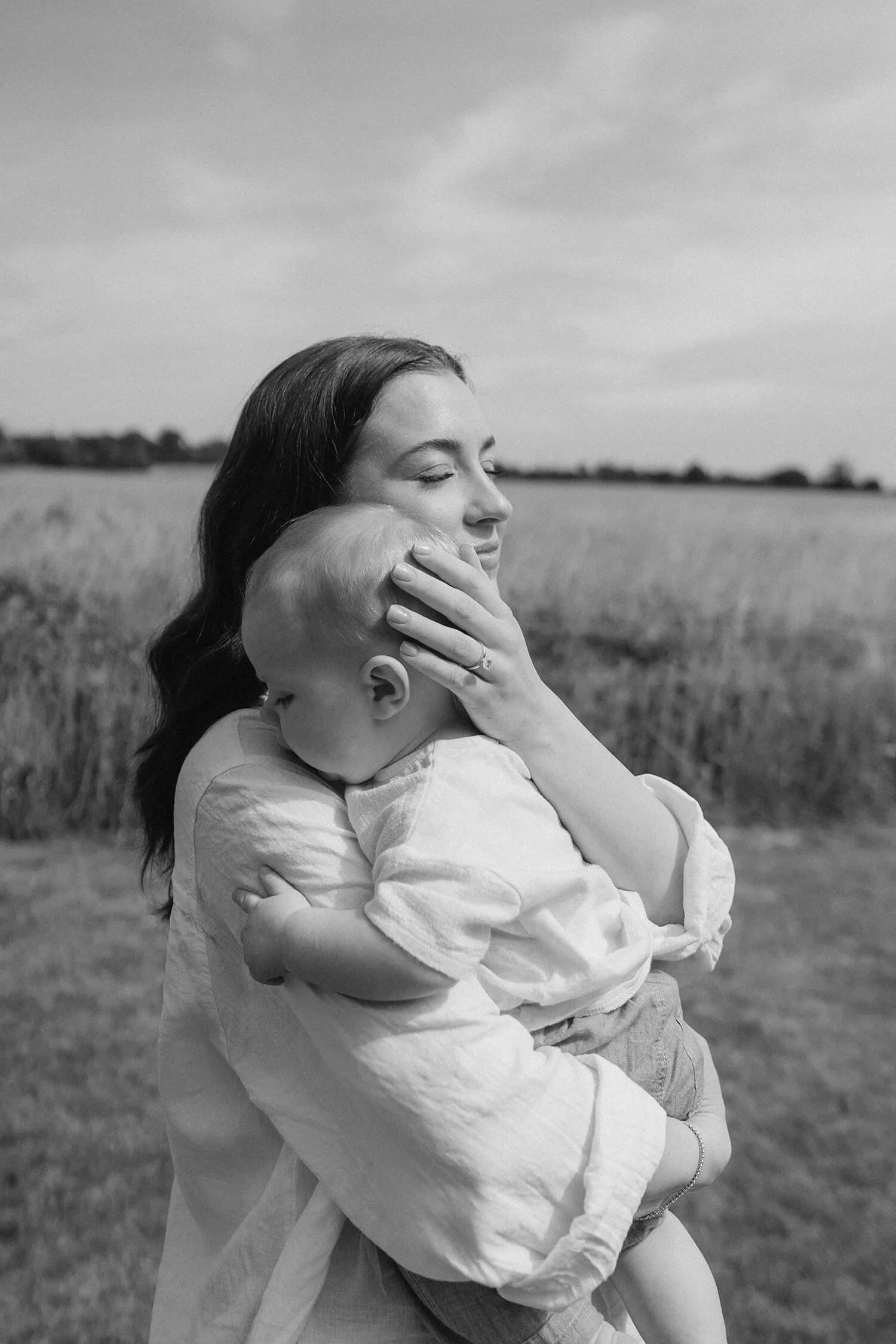 A woman holding a young child in an outdoor field, embracing and comforting them with eyes closed and a peaceful expression.