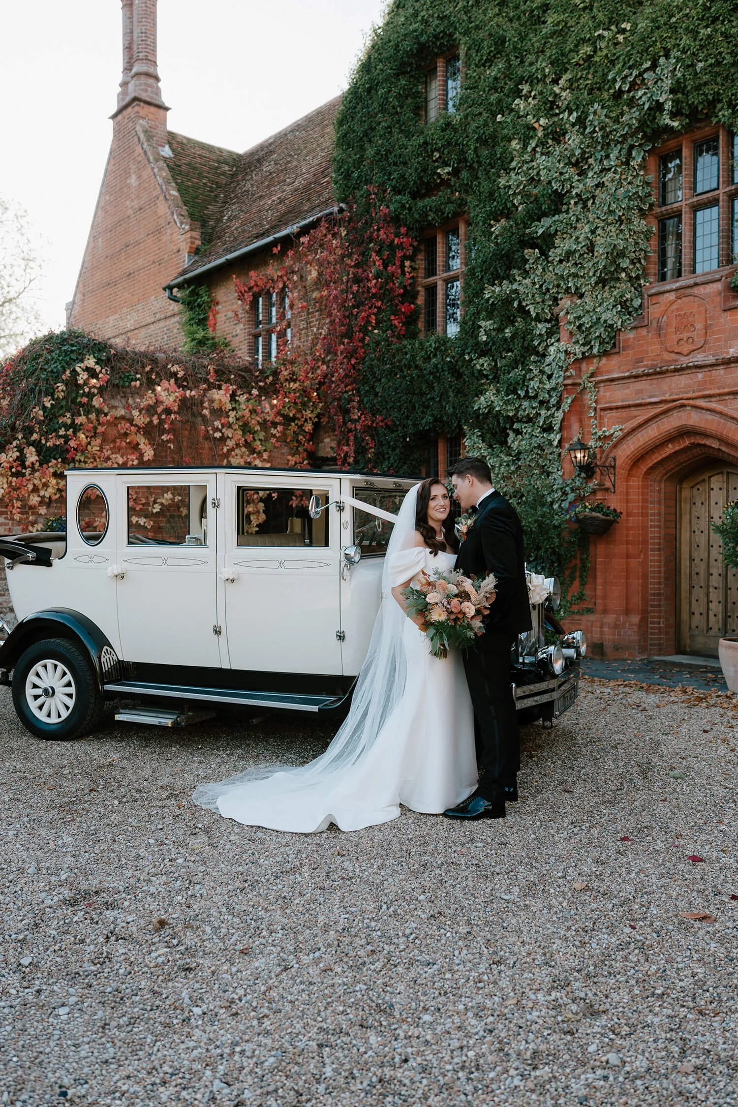 A bride and groom standing in front of a vintage white car outside a red brick building with ivy and flowers, celebrating their wedding.