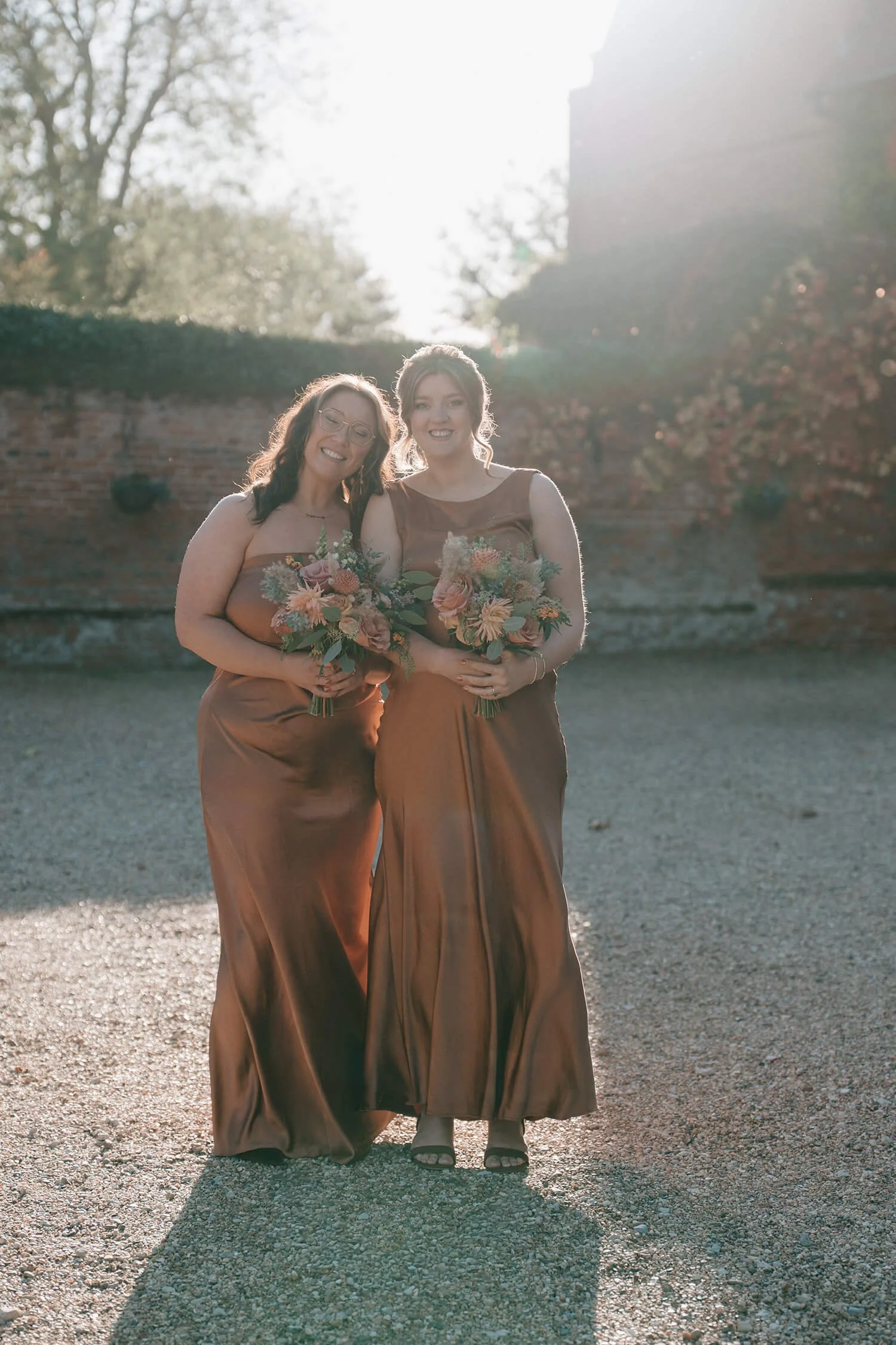 Two women in matching brown satin dresses holding flower bouquets, standing outdoors in sunlight with trees and brick wall background.
