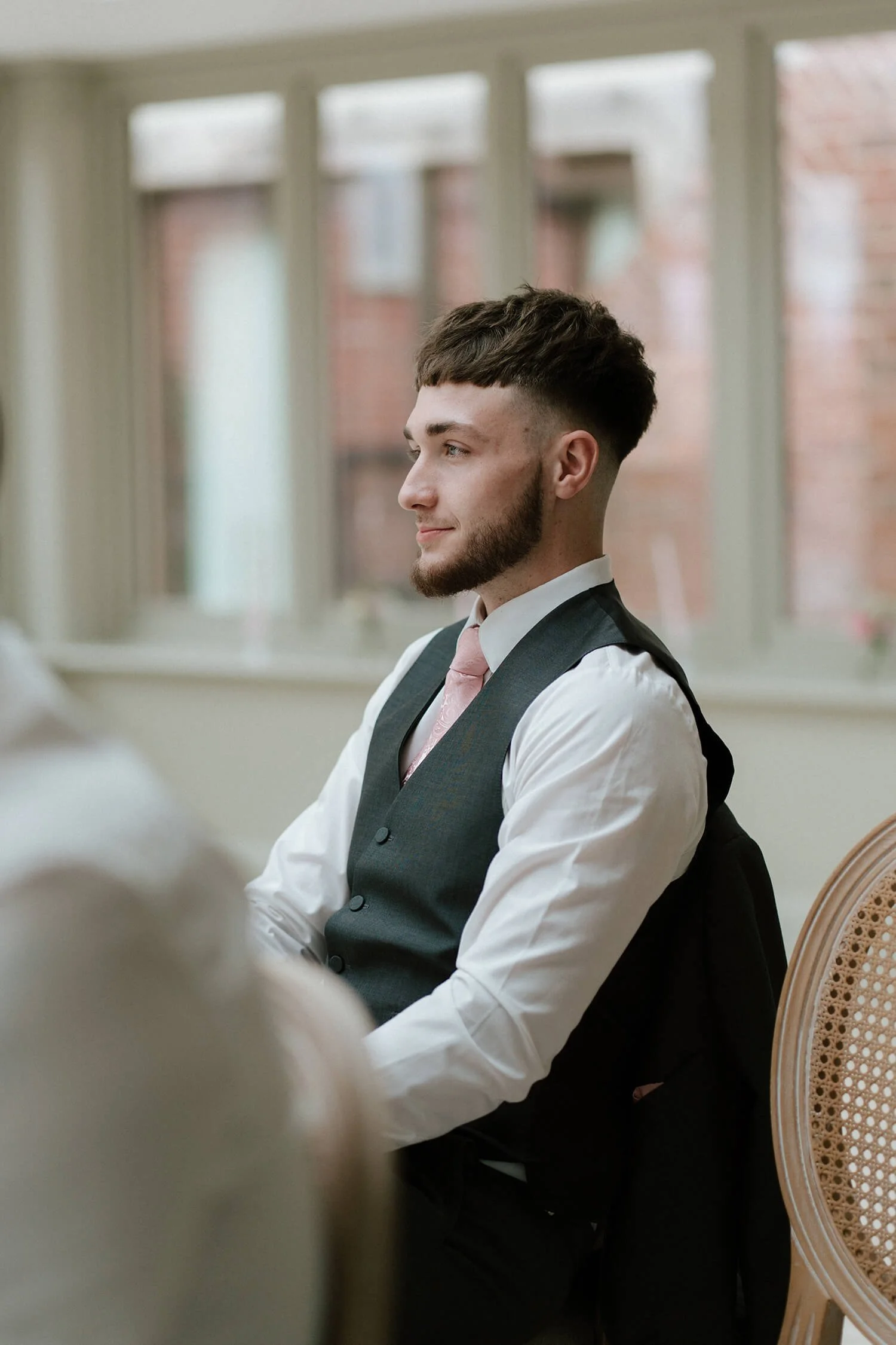 Profile view of a young man in formal attire, sitting in a room with large windows, wearing a white shirt, pink tie, and dark vest.