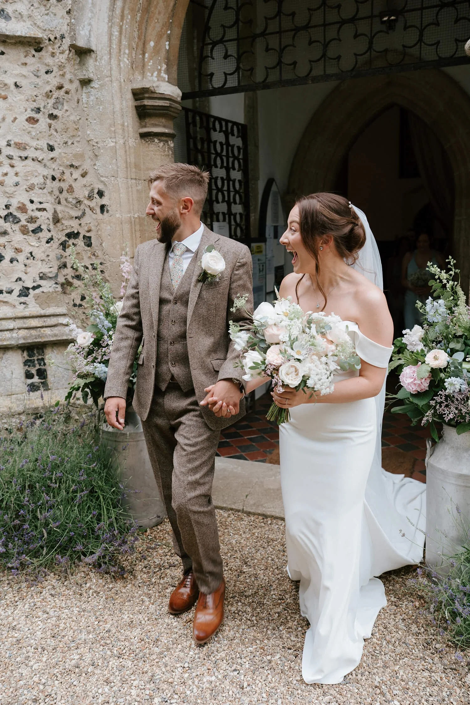 A newlywed couple walking hand in hand outside a church, smiling and laughing. The bride is in a white off-shoulder wedding dress holding a bouquet of white and pink flowers. The groom is in a brown tweed suit with a white boutonnière, wearing brown 