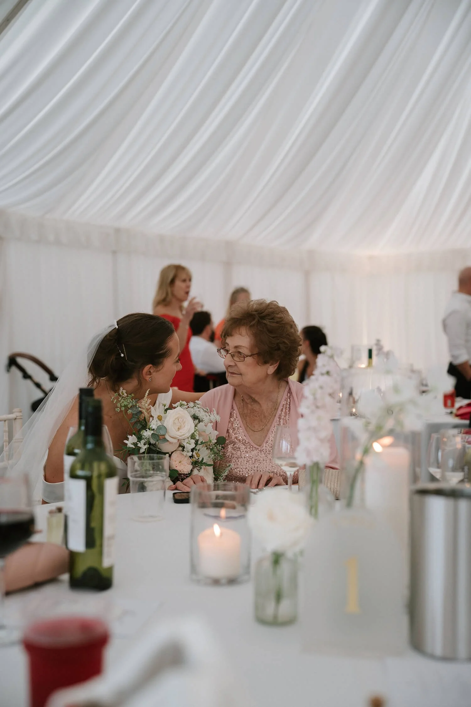 A bride and an elderly woman, possibly her grandmother, sit at a wedding reception table decorated with candles and flowers, in a bright tent-style setting.