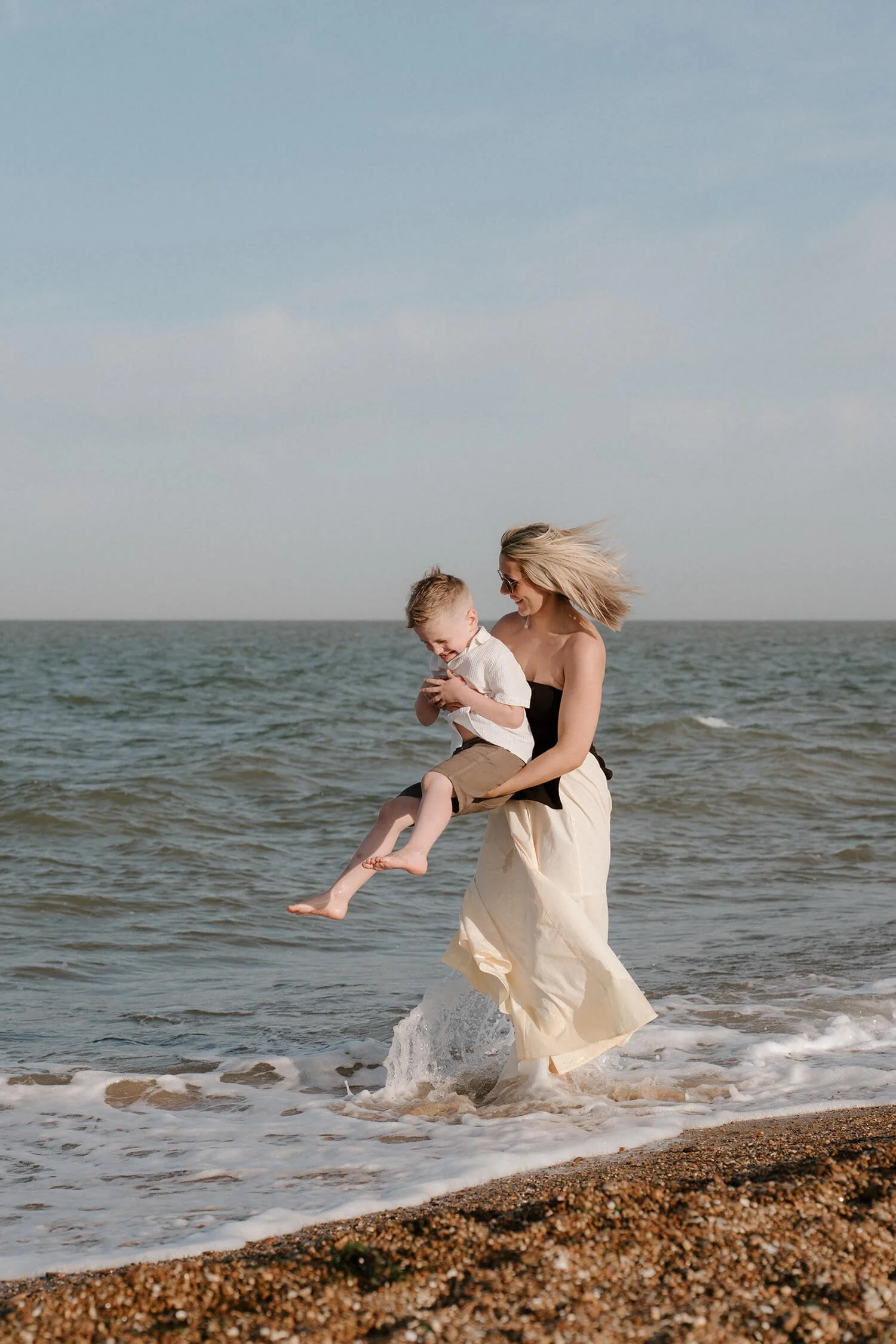 A woman holding a young boy in her arms as they play near the ocean shoreline, with waves and a cloudy sky in the background.