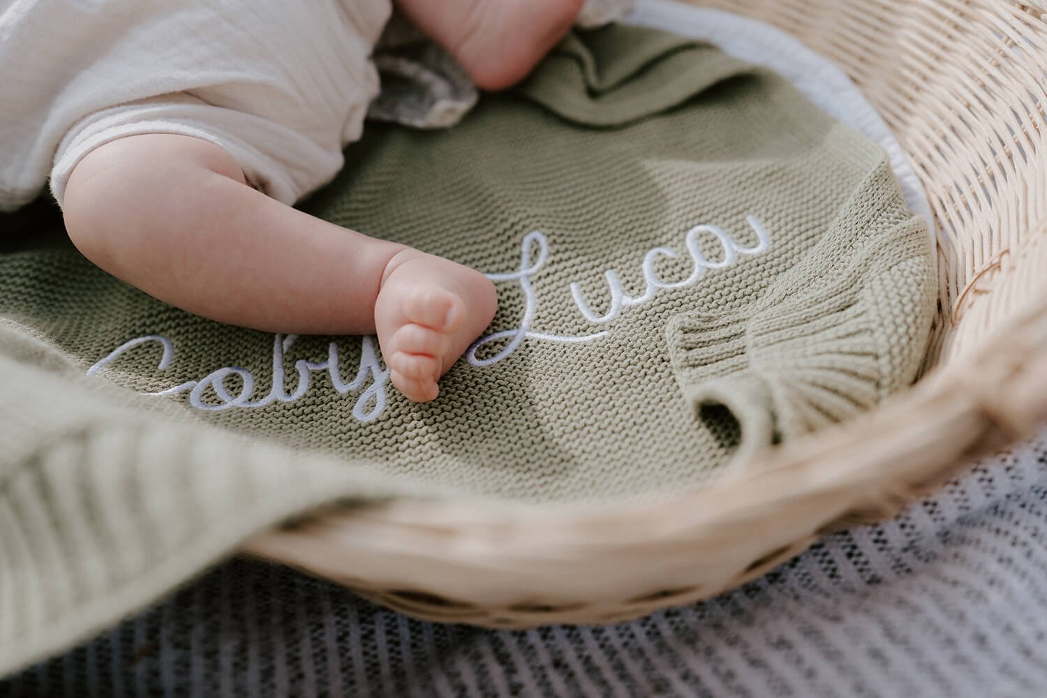 Close-up of a newborn baby’s hand resting on a beige knitted blanket with the embroidered name 'baby lucas' in white thread, inside a wicker basket.
