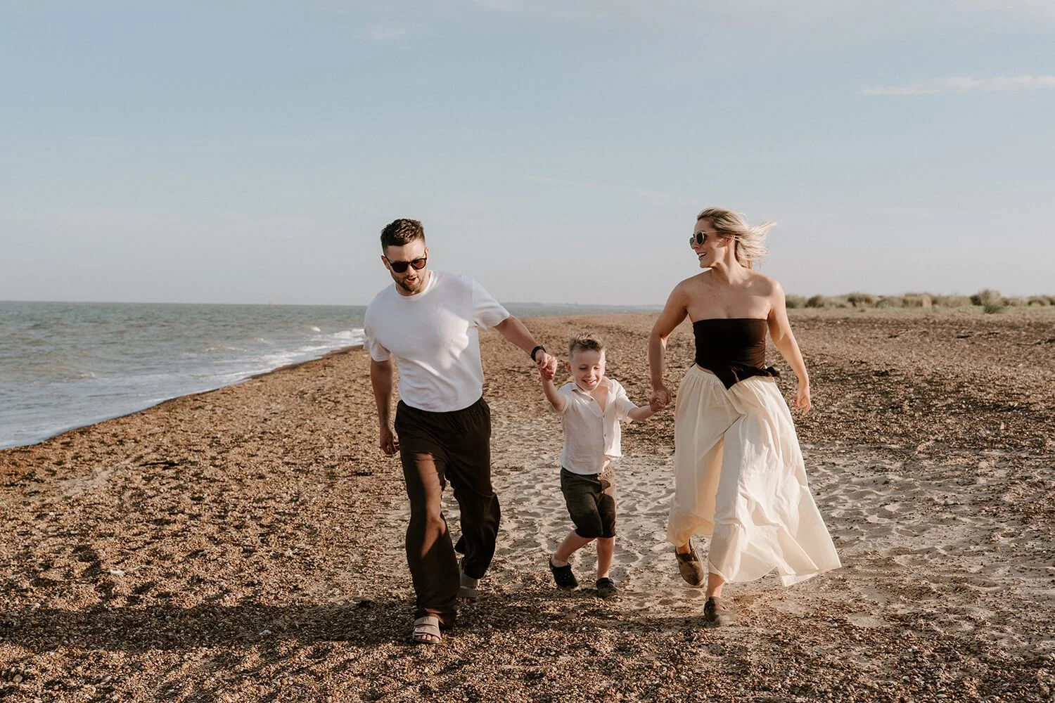 A family of three, a man, a woman, and a young boy, running hand in hand along a sandy beach with the ocean in the background. The man is wearing a white t-shirt and black pants, the woman is wearing a strapless top and a long skirt, and the boy is i