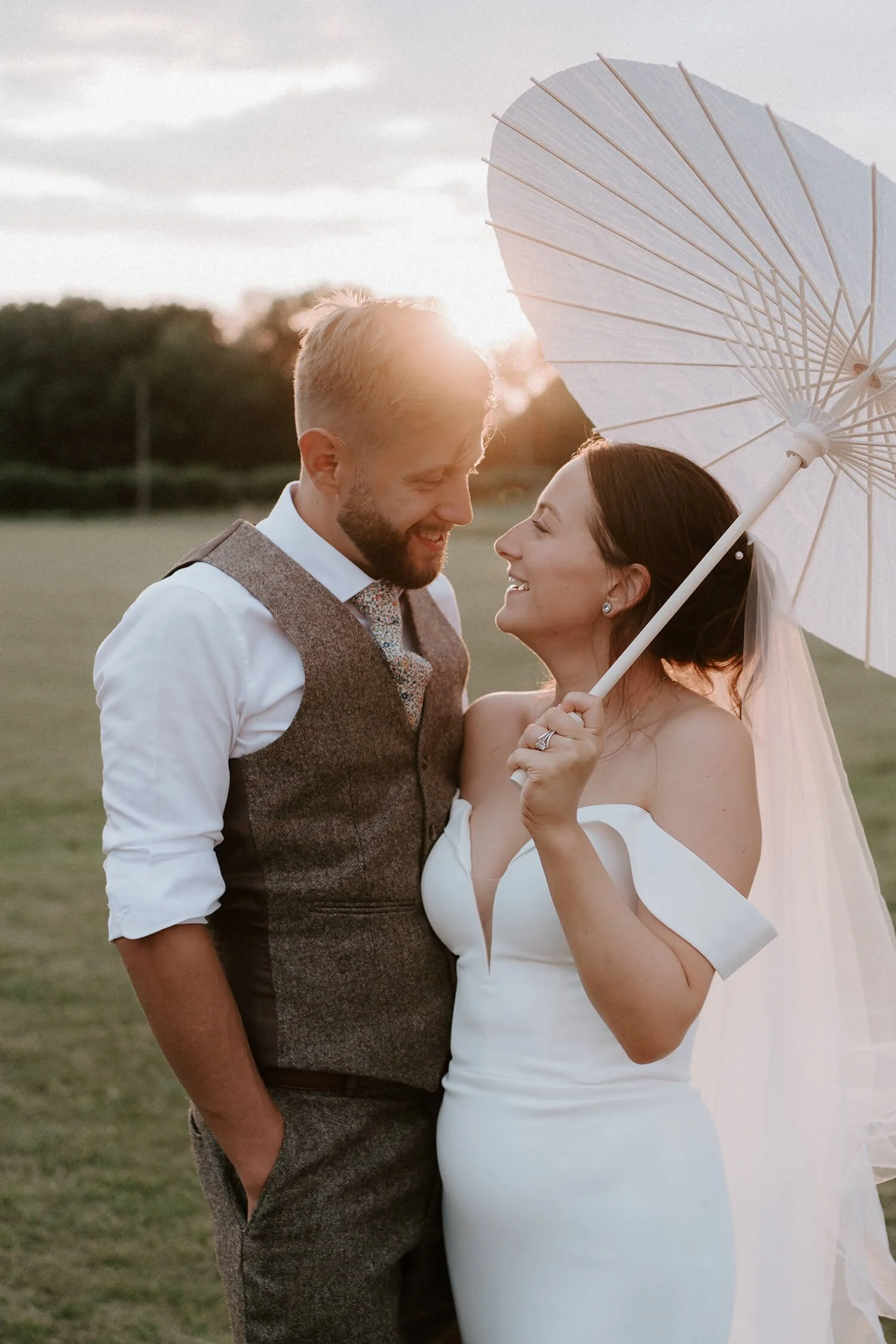 A newlywed couple smiling and looking at each other outdoors during sunset, with the bride holding a white parasol.