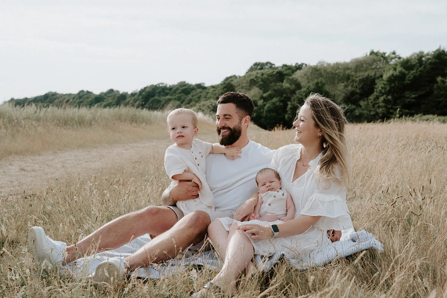 A family of four sitting on a blanket in a field of tall grass, with trees in the background, enjoying a sunny day.