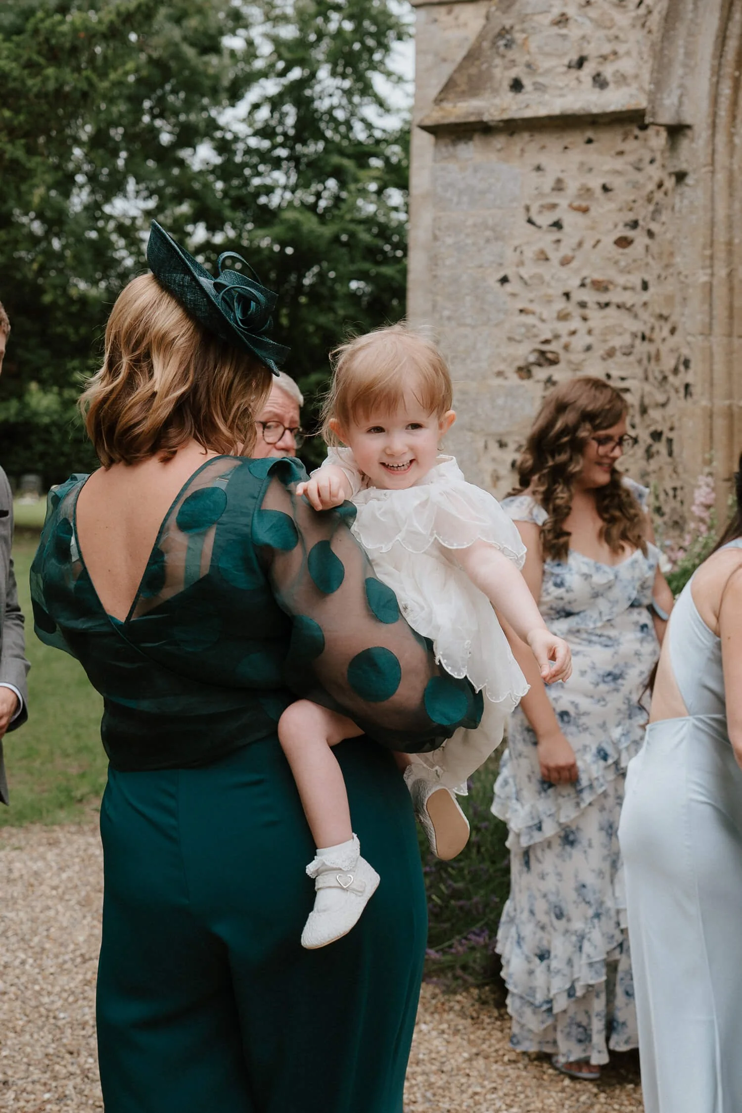 A woman holding a laughing young girl wearing a white dress at an outdoor event near an old stone building.