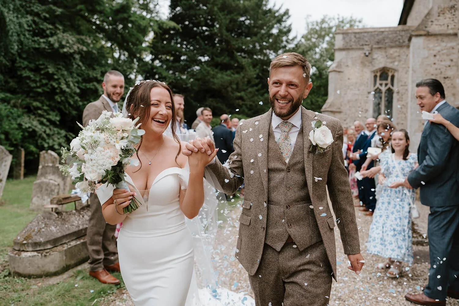 A newly married couple smiling and holding hands as they walk outdoors, surrounded by friends and family throwing confetti during a wedding celebration.