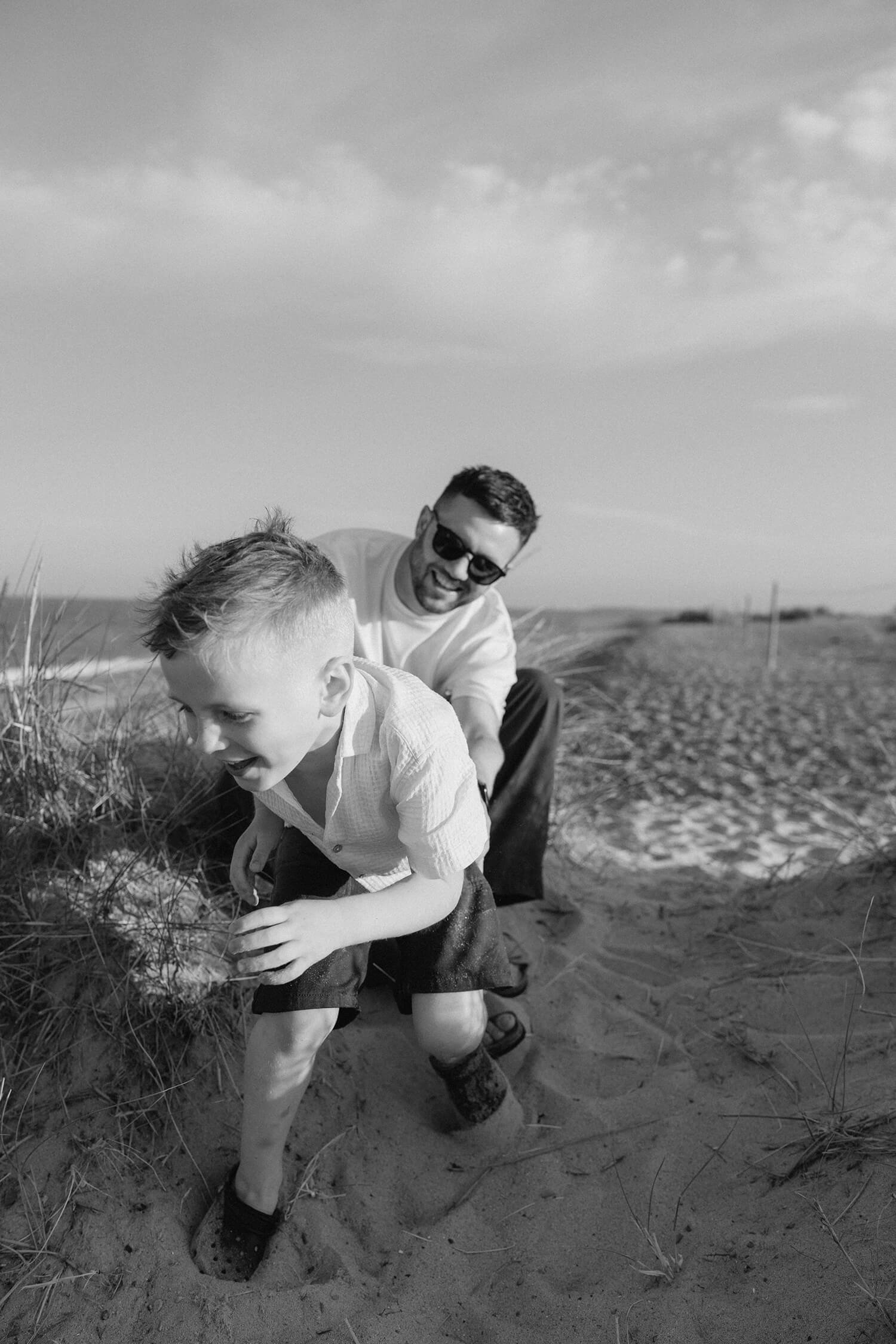 A black and white photo of a man and a young boy on a beach, crouching near sand dunes. The man is wearing sunglasses and a white shirt, smiling at the boy. The boy has light-colored hair, is wearing a light shirt and shorts, and appears to be explor