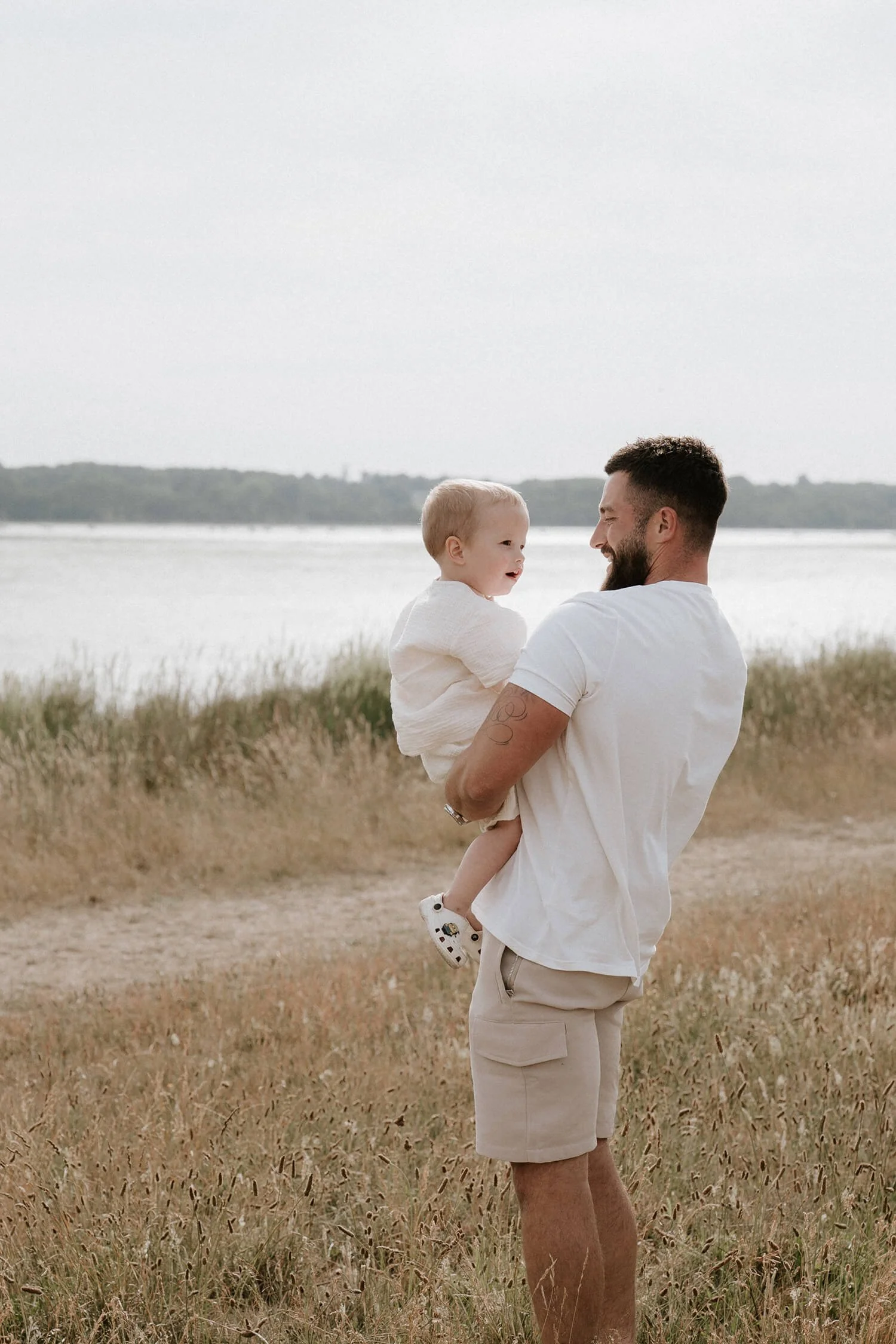 A man holding a young child outdoors near a body of water with grassy shoreline, both smiling.