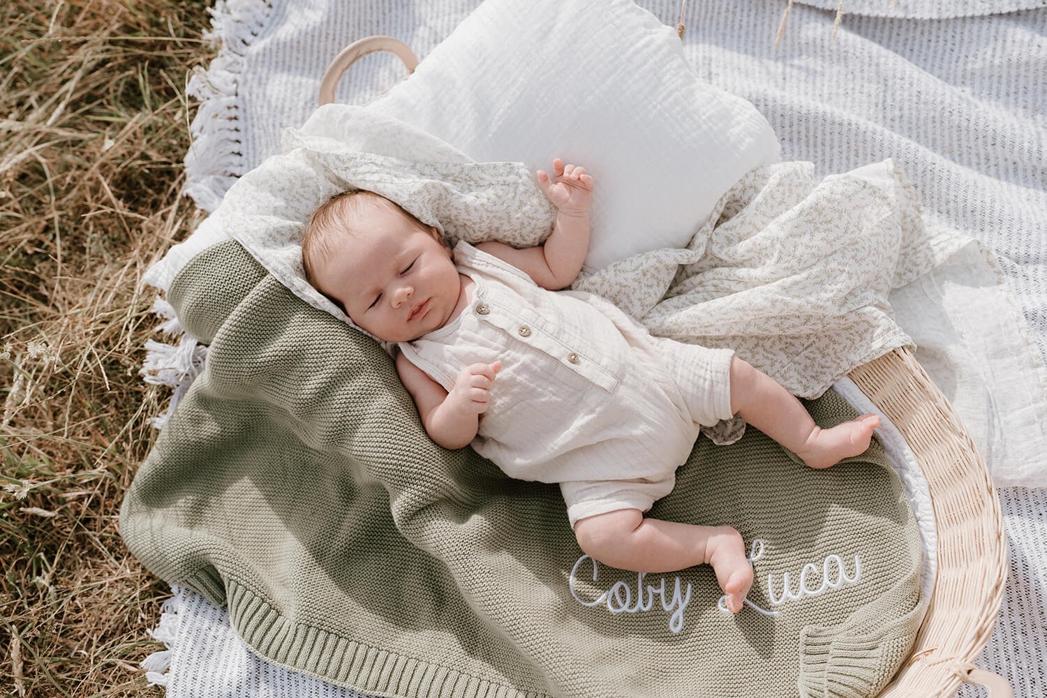 A sleepy baby lying on a green blanket inside a wicker basket, with grass surrounding the basket.
