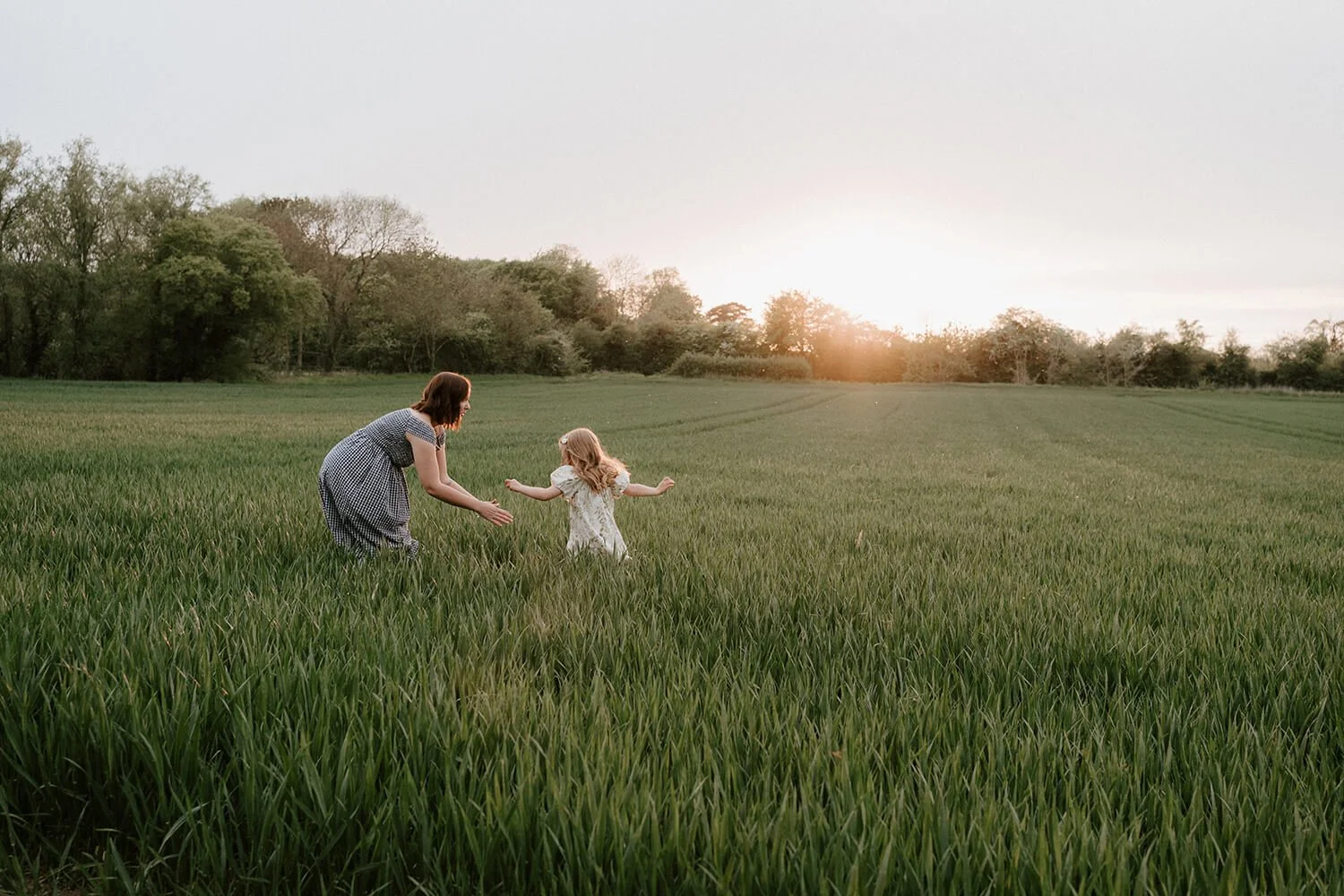 A woman and a young girl playing in a green field at sunset, holding hands and running.