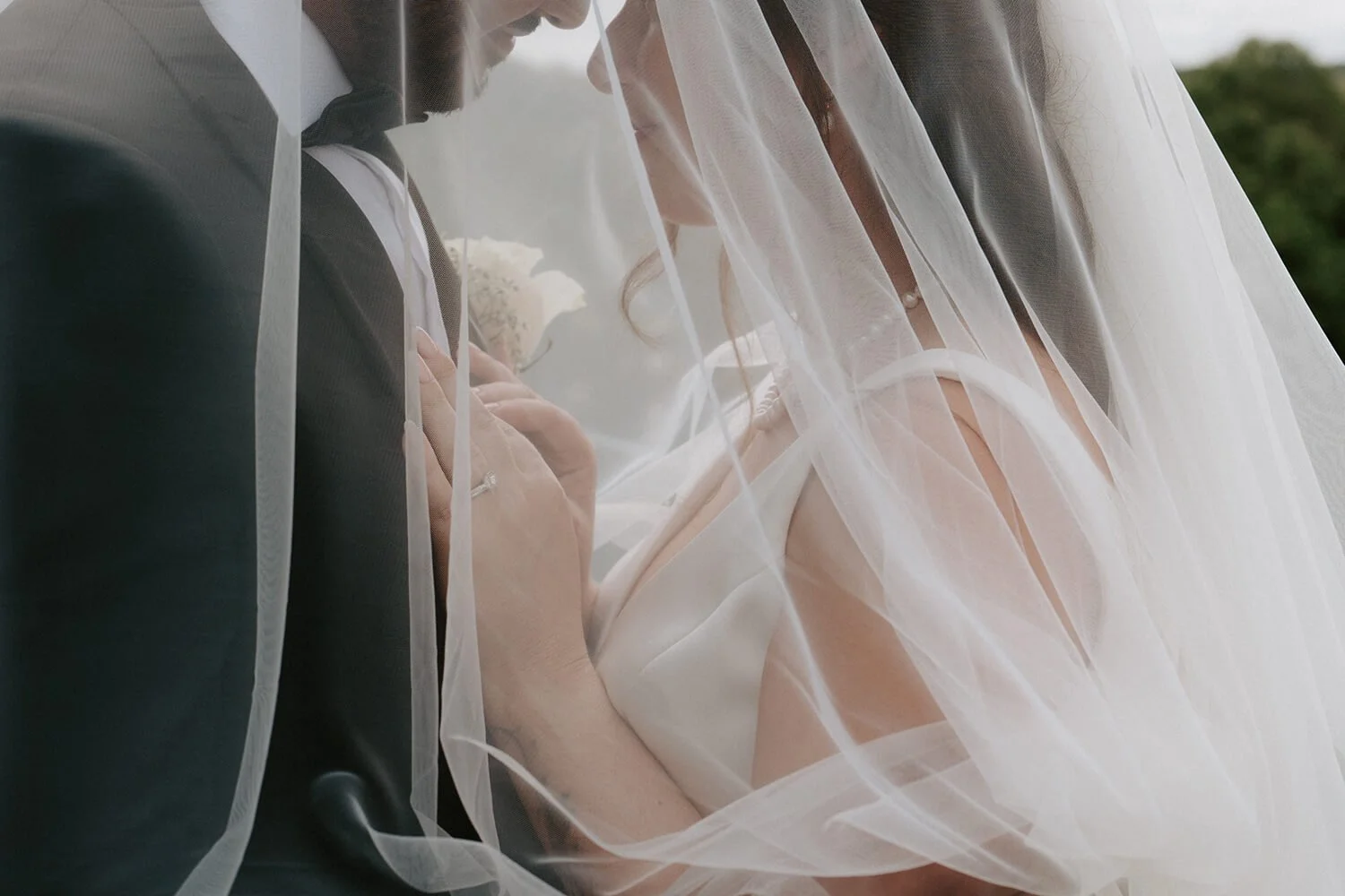 A bride and groom face each other closely, with the bride's hand on the groom's chest, both under a sheer white veil during a wedding ceremony.