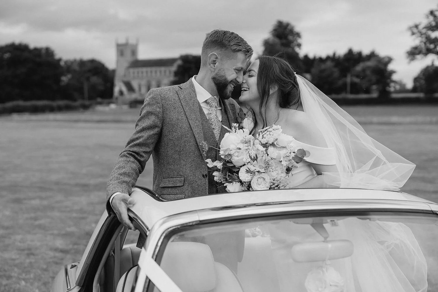 Black and white photo of a newlywed couple smiling and leaning noses together, standing next to a vintage car with a bouquet of flowers, in an outdoor grassy area with trees and a church in the background.