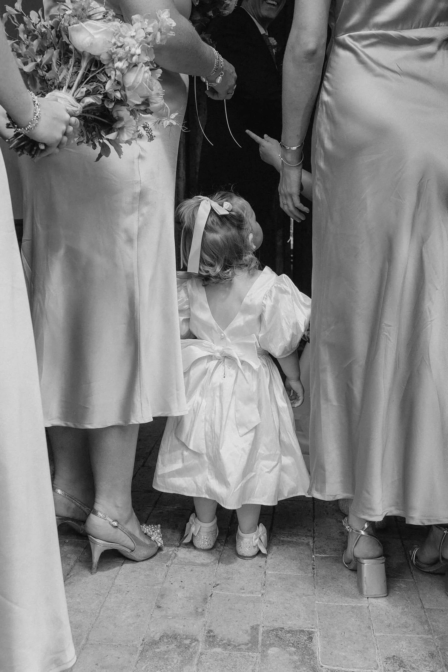 A young girl in a white dress with a bow in her hair is standing between two women, holding their hands, at what appears to be a wedding ceremony. The women are wearing elegant dresses and heels, and one is holding a bouquet of flowers.