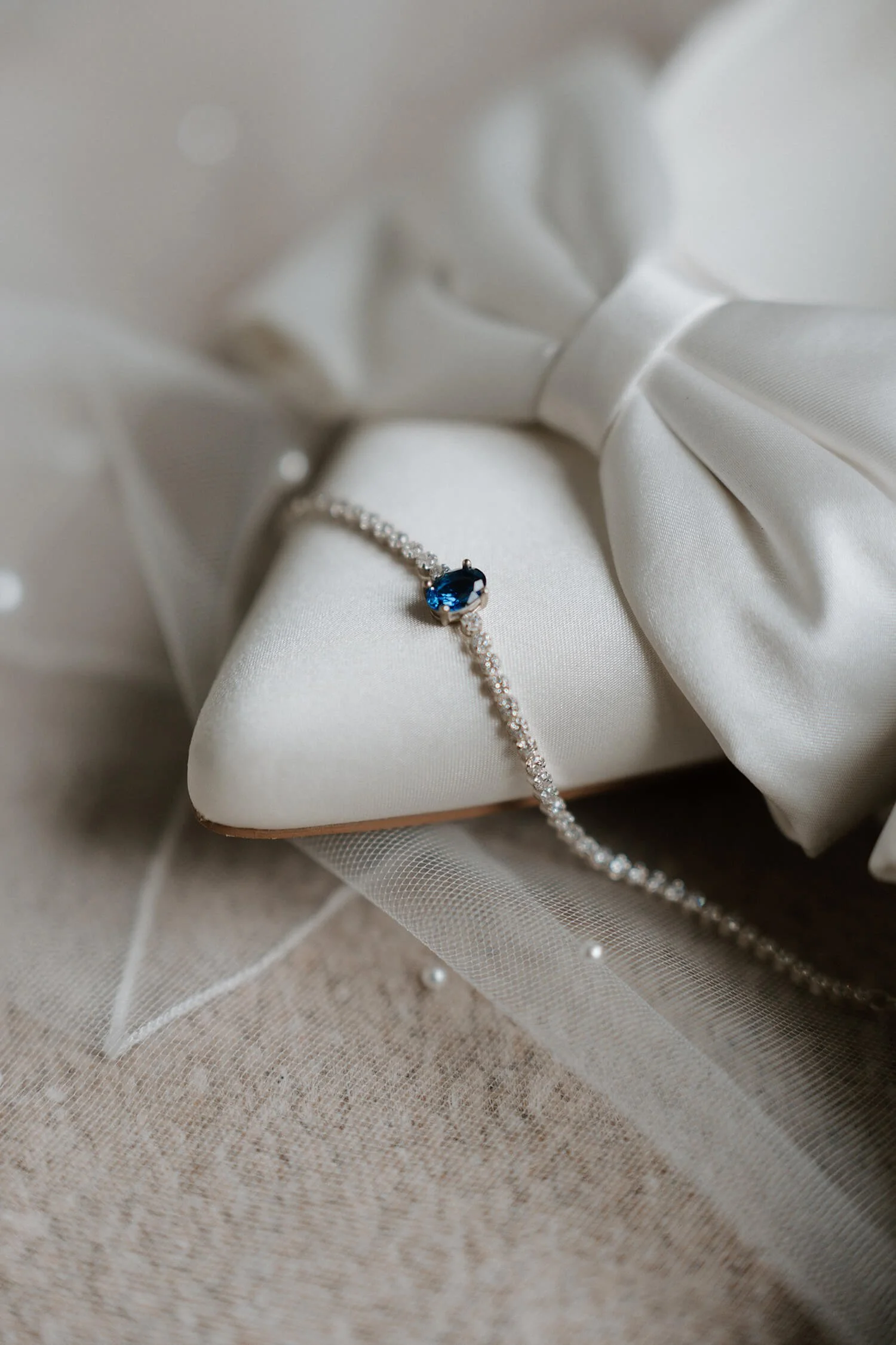 Close-up of a diamond necklace with a blue gemstone, resting on a white satin object decorated with a bow, possibly a jewelry box.