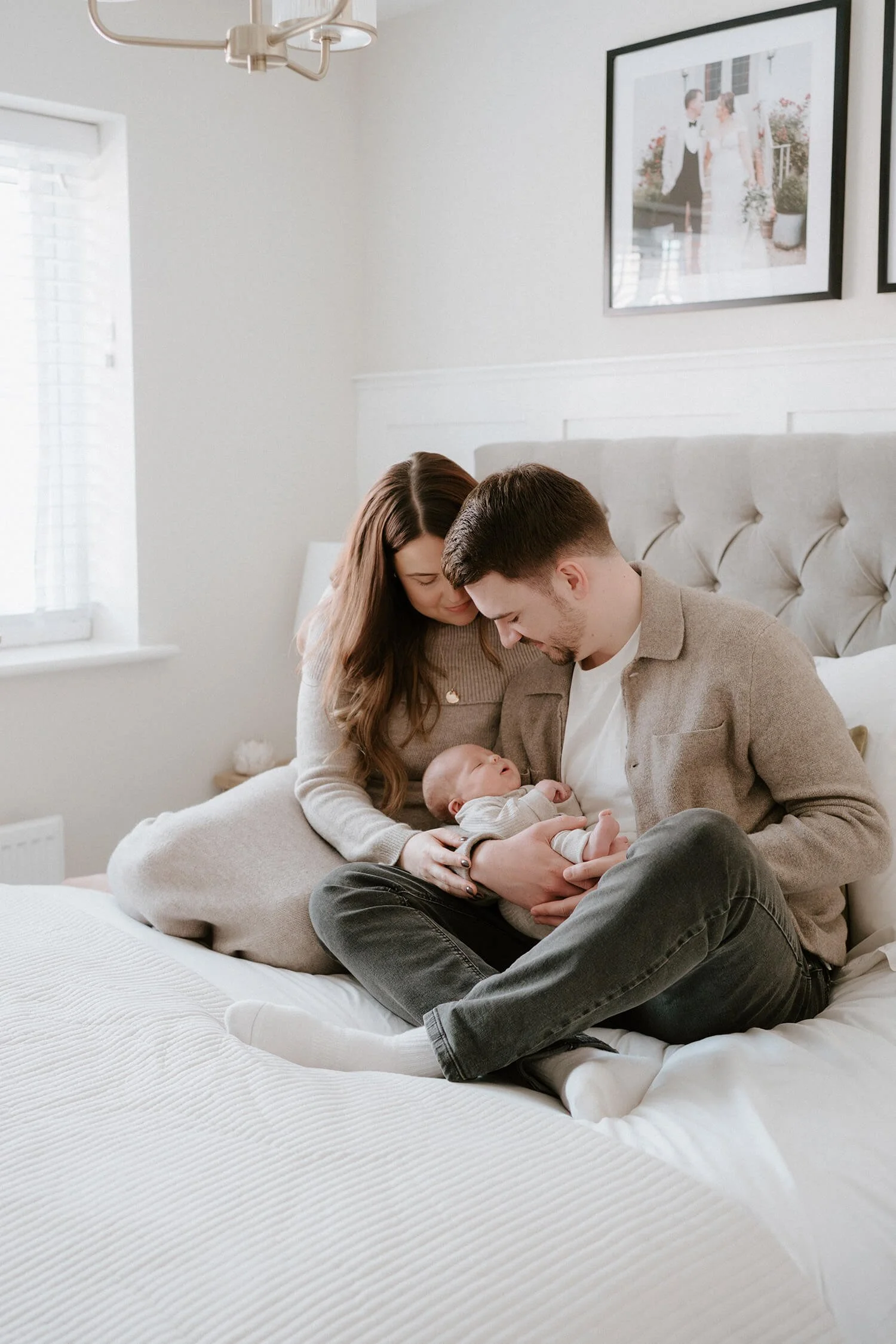 A family of three sitting on a bed in a cozy, well-lit bedroom. The mother and father are gently holding their newborn baby, looking at the baby with affection. The room features white walls, a tufted headboard, framed photos on the wall, and natural light coming through a window.