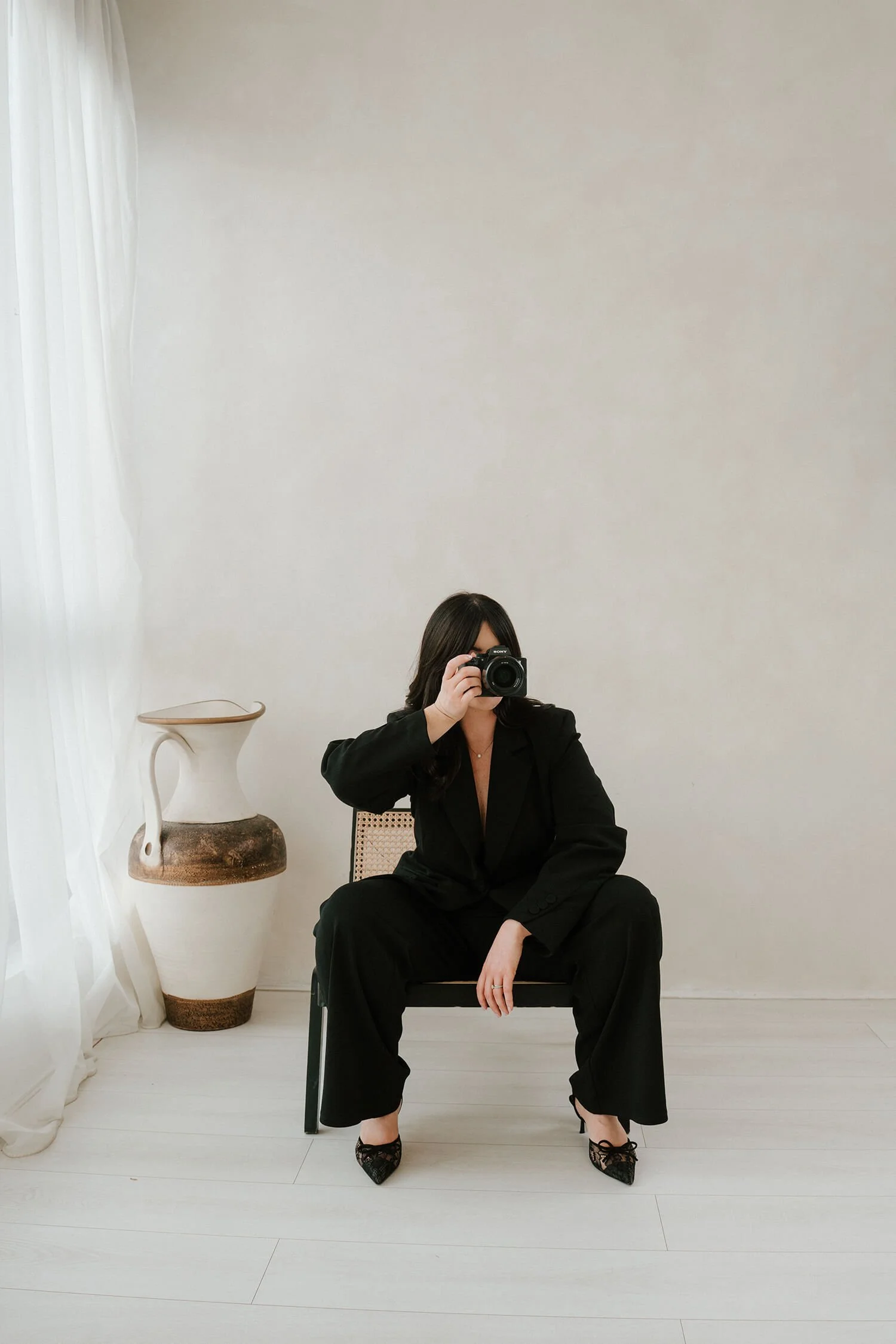 Woman in black suit sitting on a chair, taking a photo of herself in a mirror, with a large ceramic vase to her side