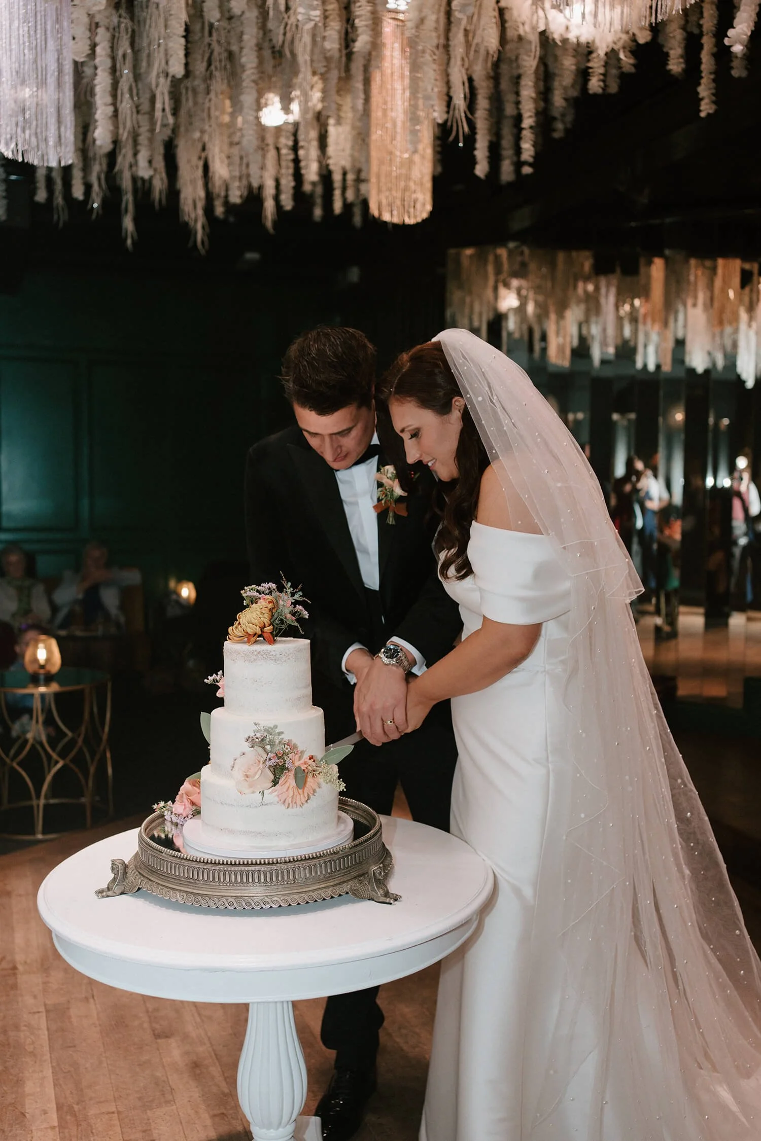 Bride and groom cutting a wedding cake together indoors with hanging decorations and guests in the background.