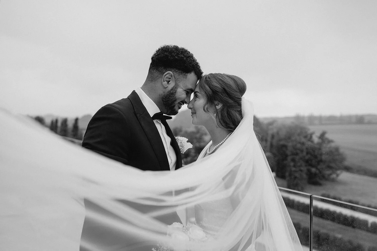 Black and white photo of a bride and groom with their foreheads touching, smiling. The groom is wearing a tuxedo, and the bride is in a wedding dress with a veil and pearl necklace. They are outdoors with a rural landscape in the background.