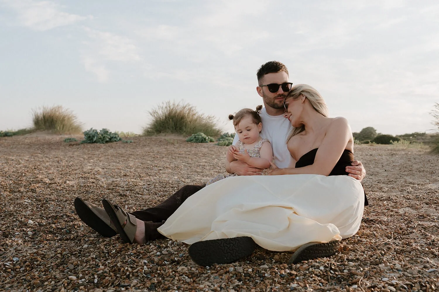 A family of three sitting together on rocky ground outdoors, with bushes and a cloudy sky in the background. The man is wearing sunglasses and a white shirt, the woman is in a black strapless dress, and the little girl is dressed in a floral dress.