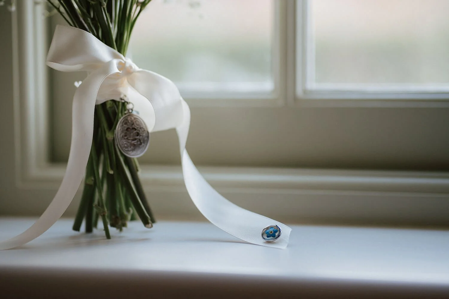 Bouquet of flowers tied with a white ribbon and decorated with a silver pendant, placed on a windowsill in front of a window.