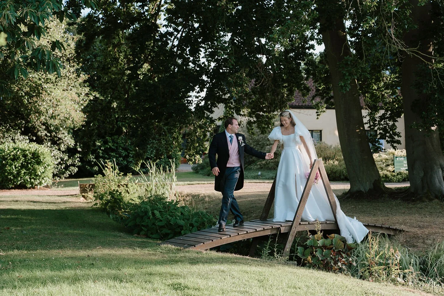 A bride and groom walking hand in hand across a small wooden bridge in a garden, surrounded by lush greenery and trees, on their wedding day.
