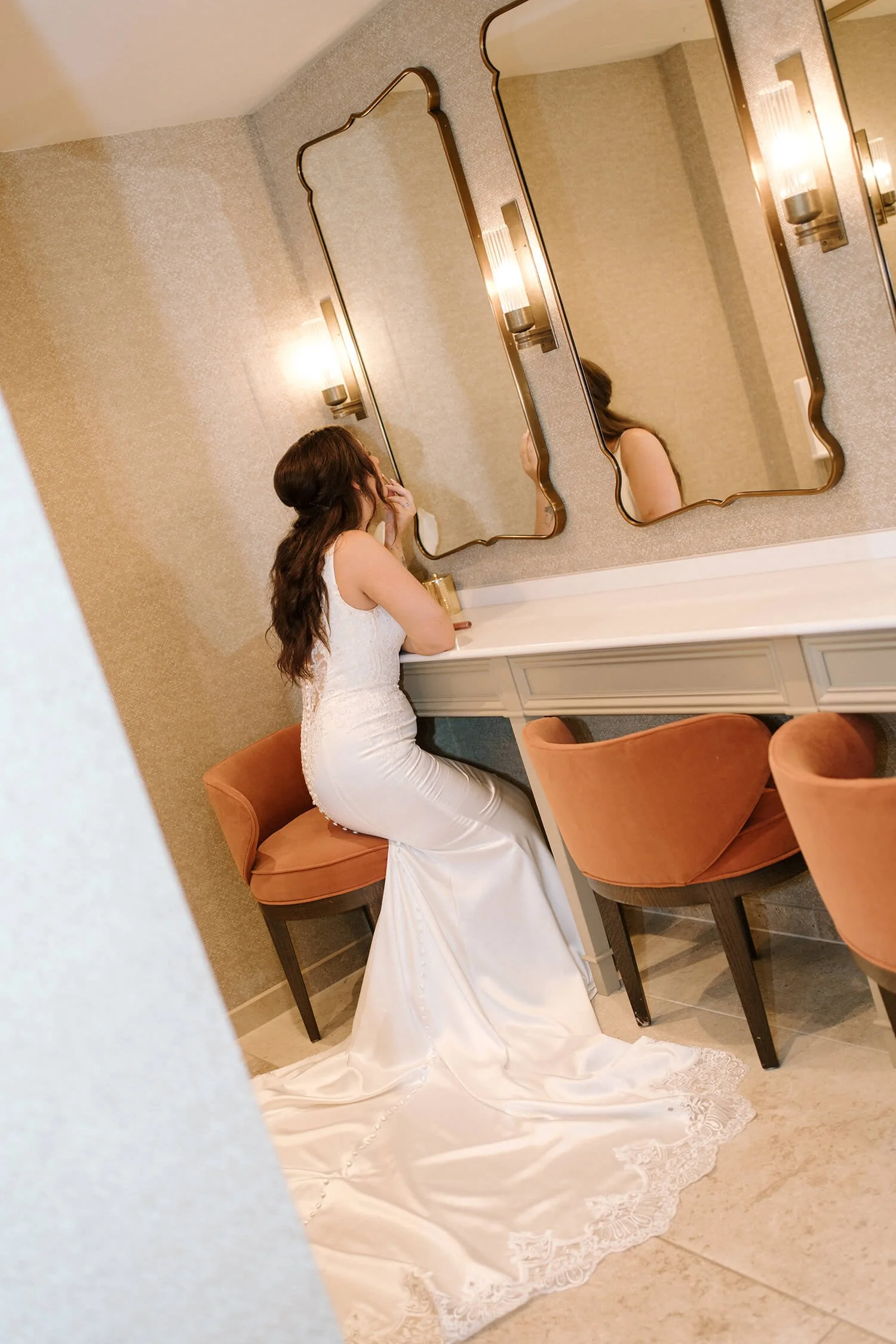 A woman in a white wedding dress is sitting on a seated vanity chair at a dressing table, looking at herself in a mirror. The room has two decorative wall mirrors, wall-mounted sconces, and beige walls.