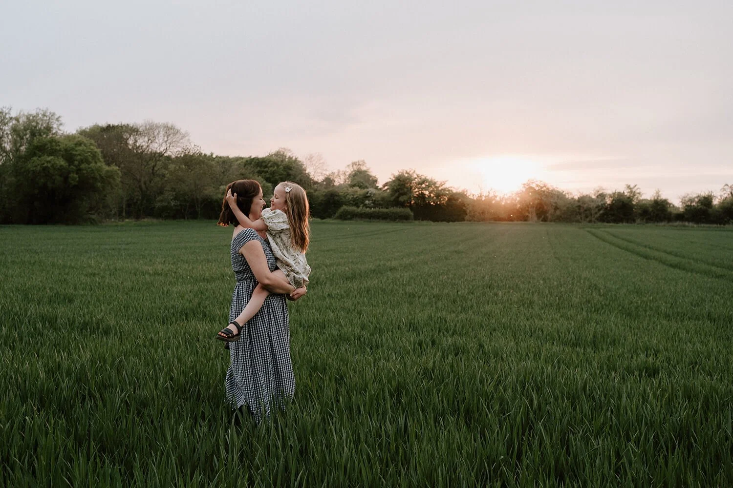 A woman holding a young girl in a grassy field at sunset, both smiling at each other.