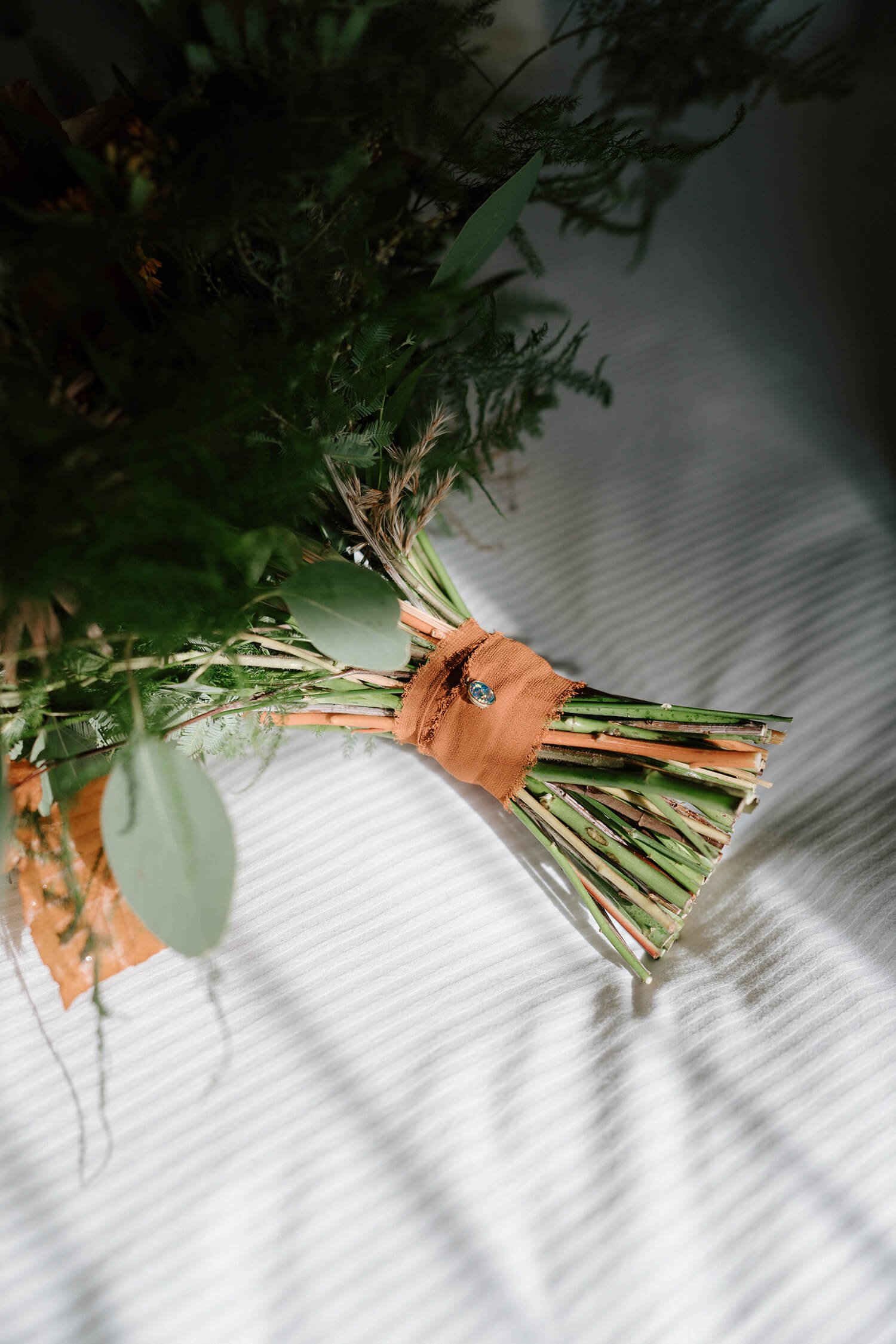 A close-up of a bouquet of assorted greenery, tied with a brown cloth ribbon with a small decorative pin, placed on a white textured surface.