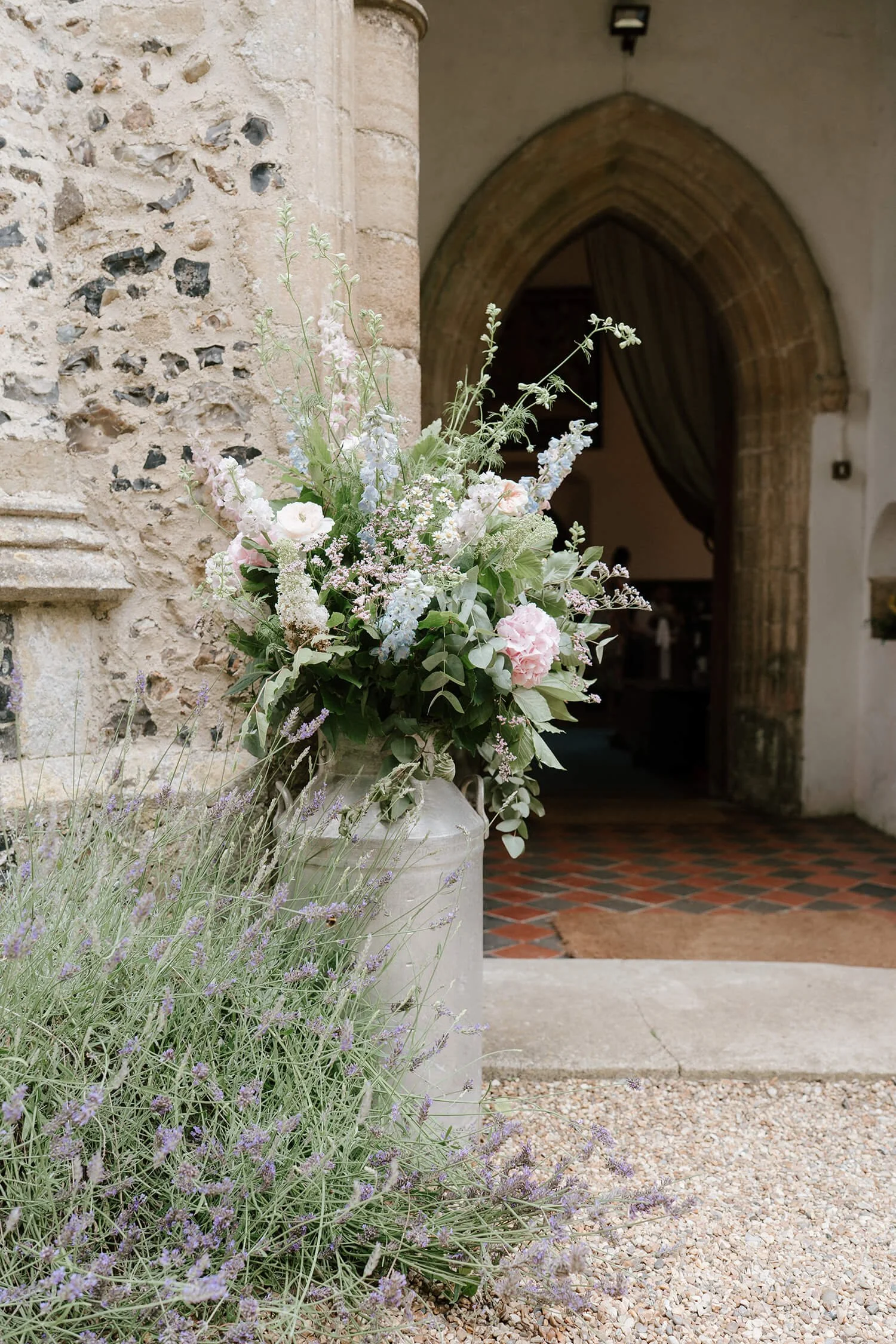Flower arrangement in a white vase with pink and white flowers and green foliage, placed outside near an old stone wall and doorway.