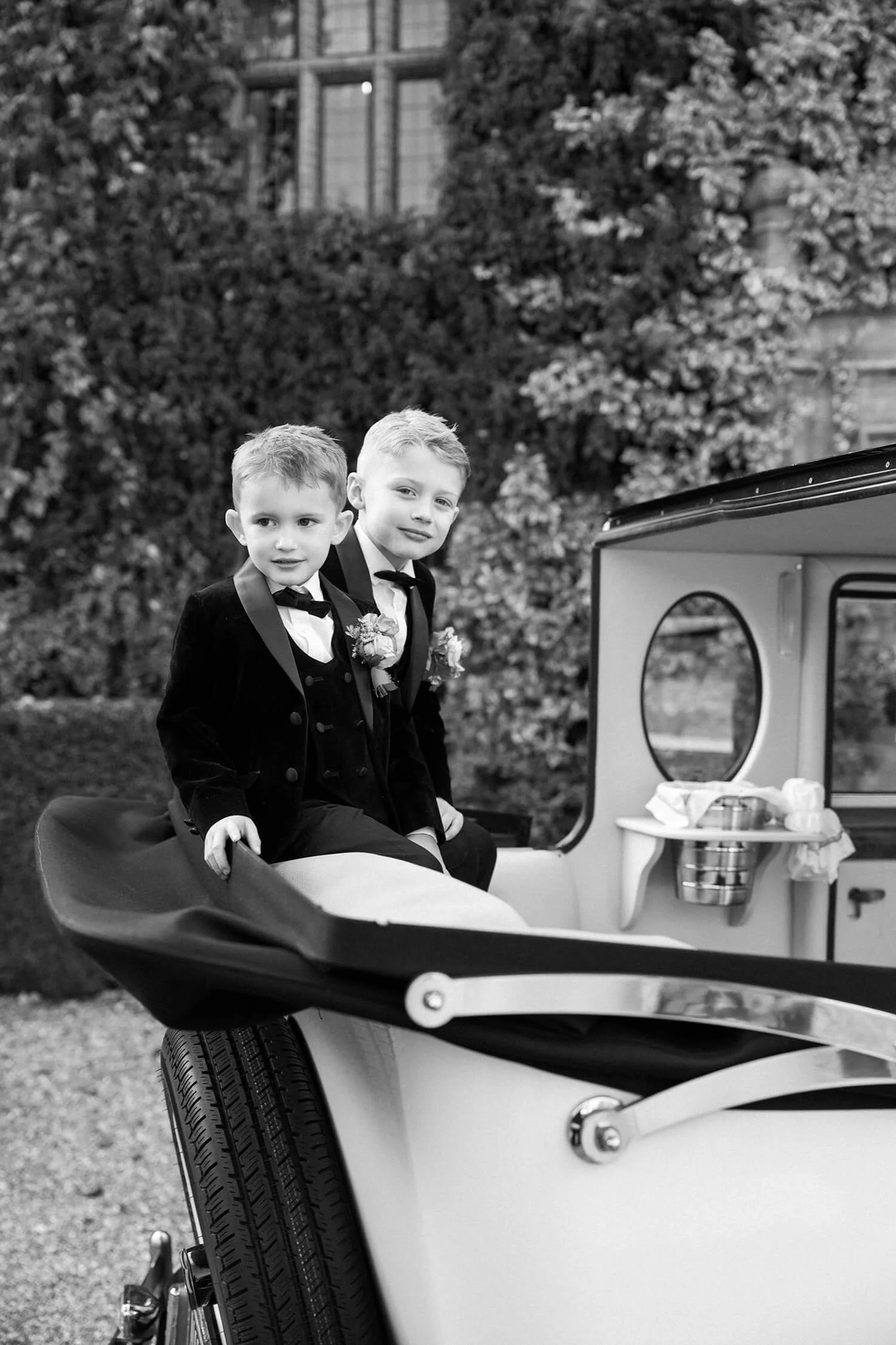 Two young boys dressed in suits and bow ties sitting on a vintage carriage, with trees and a building in the background.