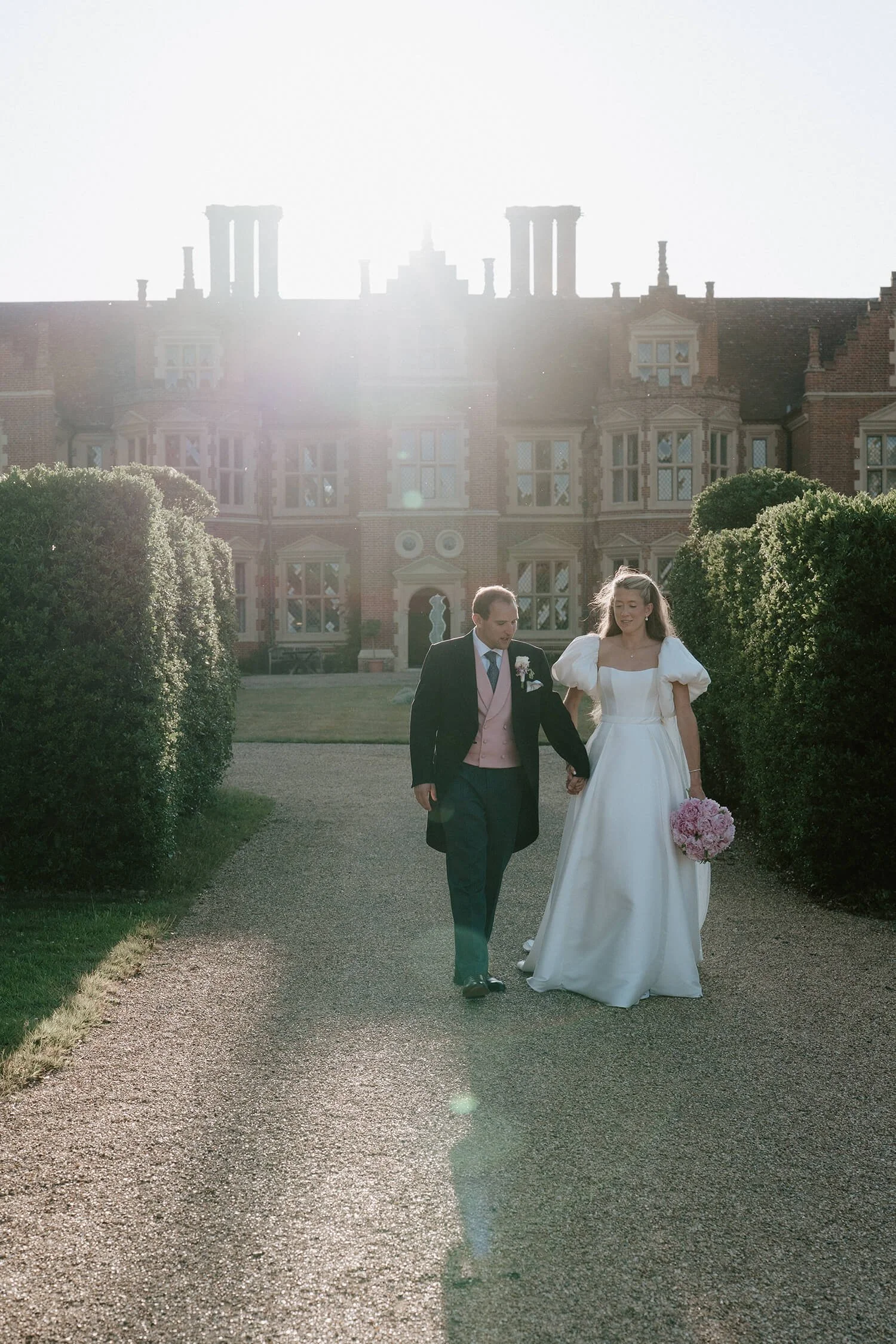 A bride and groom walking hand in hand on a gravel path outside a large, historic mansion with a garden in the background, during a wedding photoshoot in daylight.