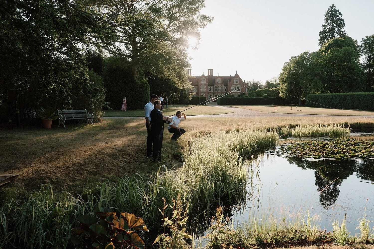 Four men standing and one man squatting by a pond in a garden, with a large mansion in the background, during sunset.
