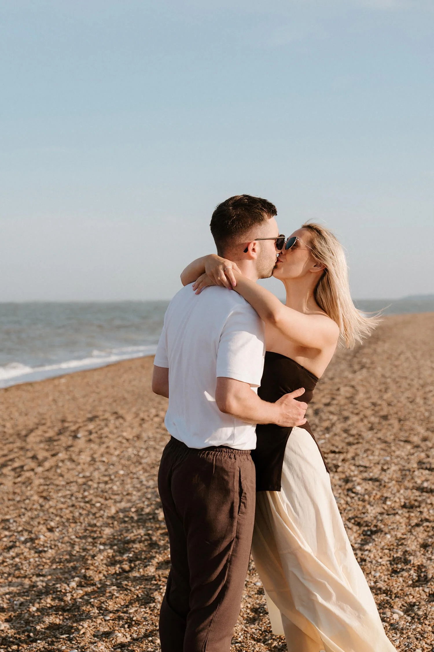 A couple kissing on a pebble beach, with the ocean and clear sky in the background.