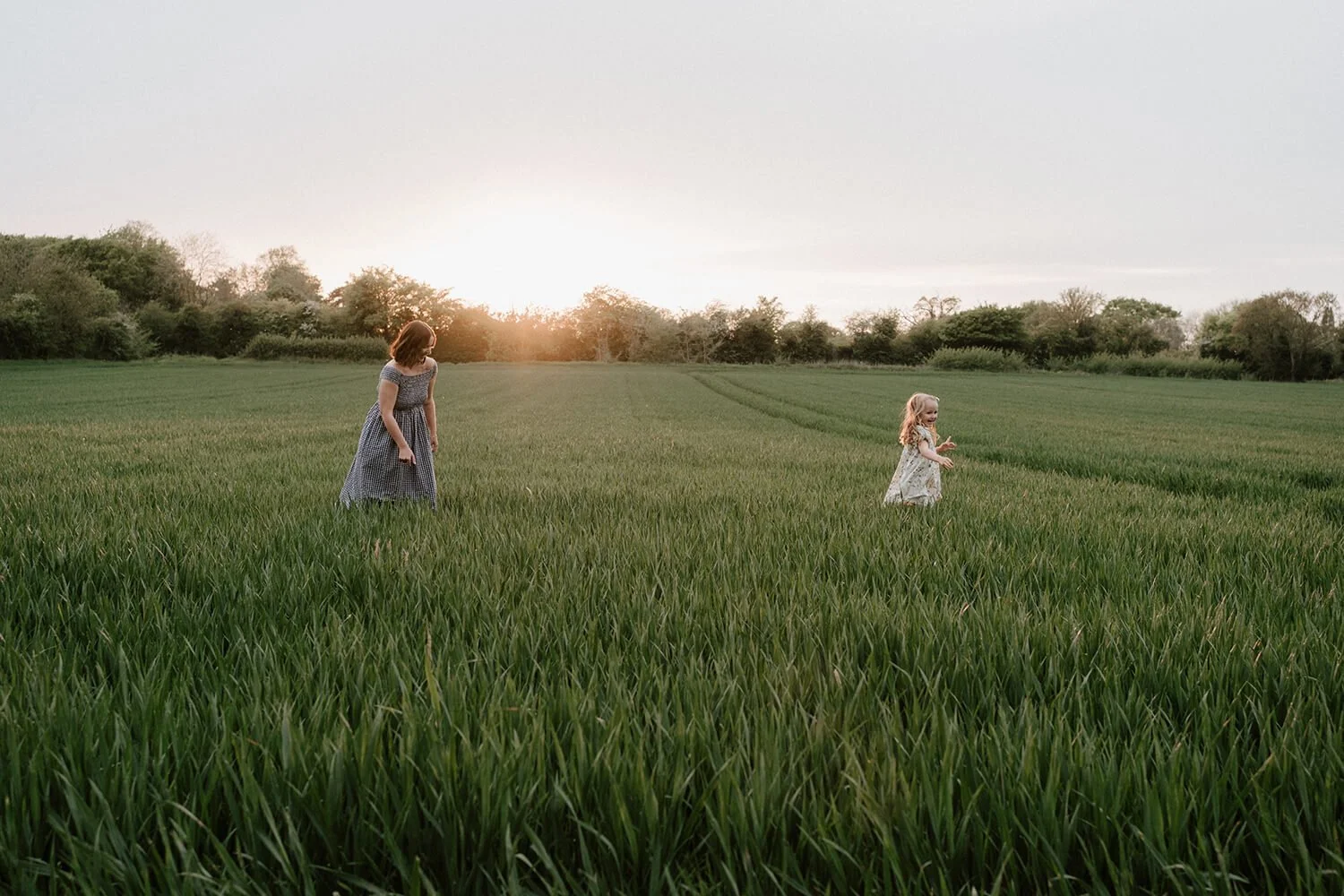 Mother and daughter walking through a green field at sunset, both wearing dresses.