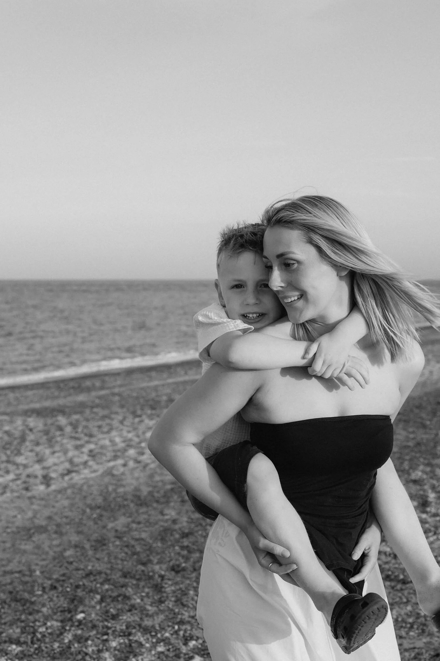 A woman holding a young boy on her back at the beach, both smiling. The woman has shoulder-length hair and is wearing a strapless top. The boy is wrapped around her neck, wearing a light shirt and shorts. The background features a calm sea and a clea